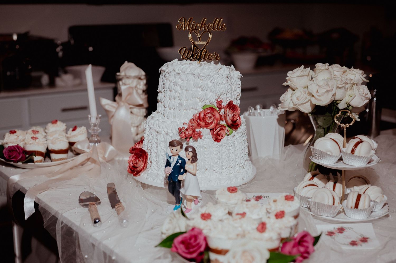 Elegant wedding cake with floral decorations and bride-groom topper on a dessert table with cupcakes and macarons.
