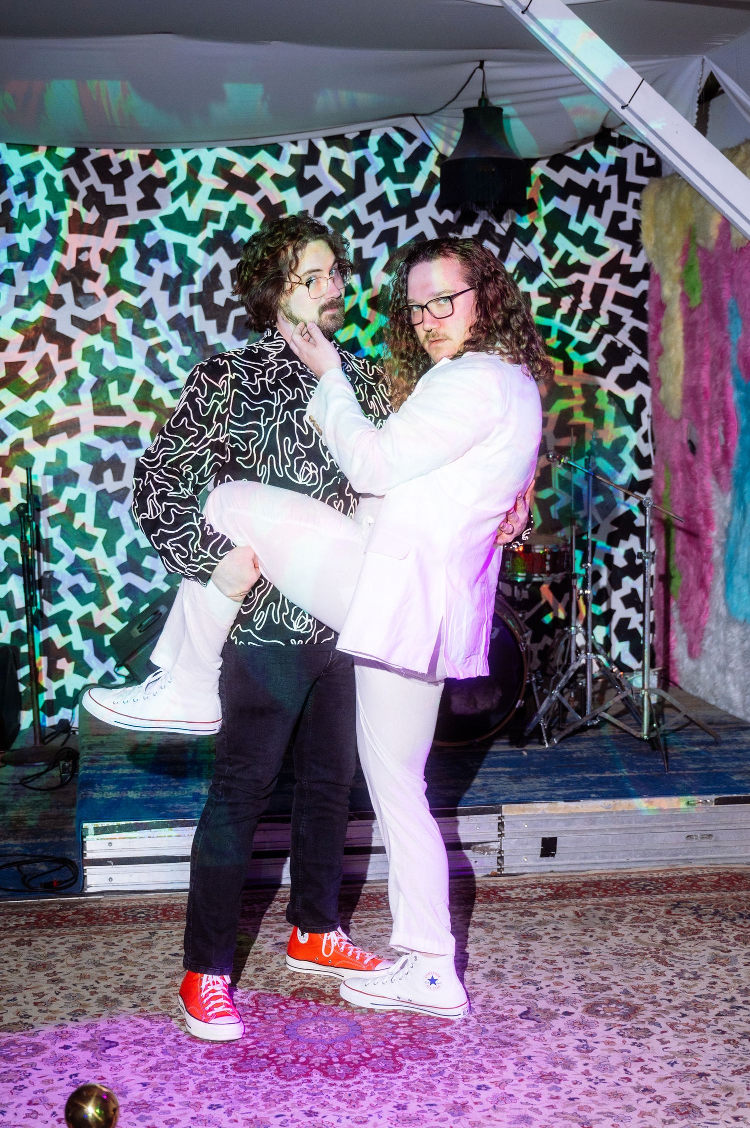 Two curly-haired people in black and white outfits pose playfully on a colorful patterned stage backdrop.