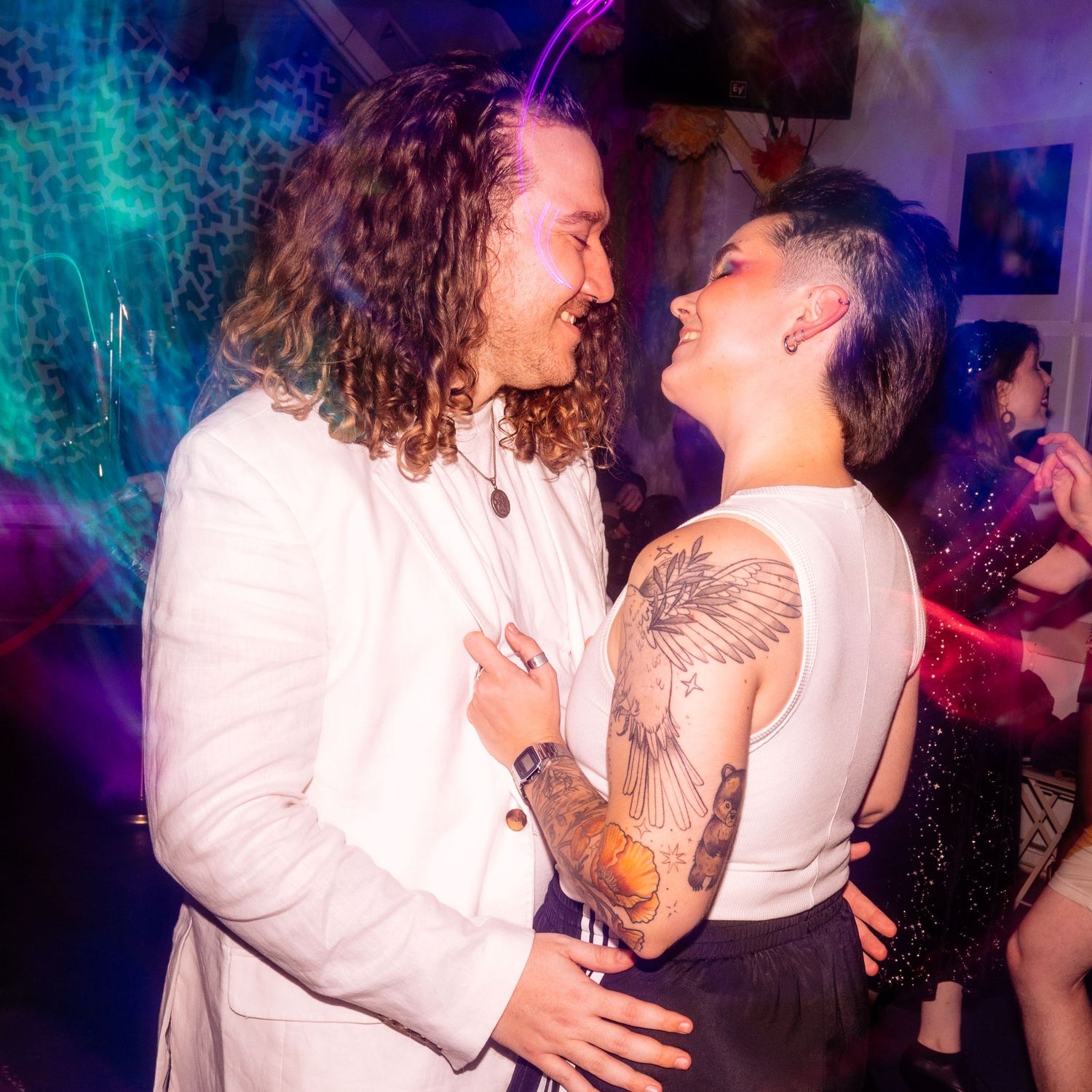 Two people sharing an intimate moment at a colorful party, one in a white blazer with curly hair, the other with tattoos and short hair.