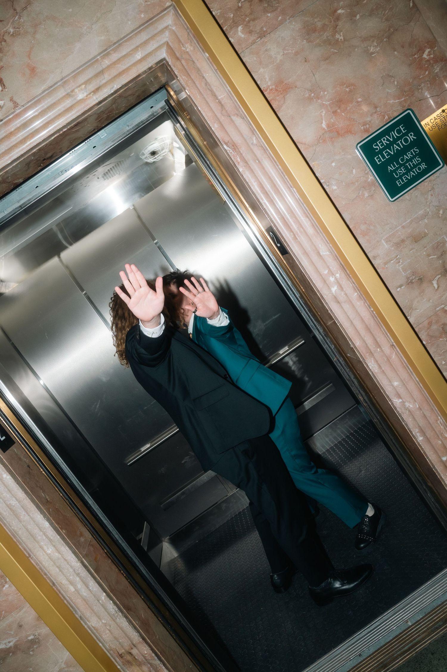 Person in teal suit shielding face with hand inside a service elevator with marble walls.