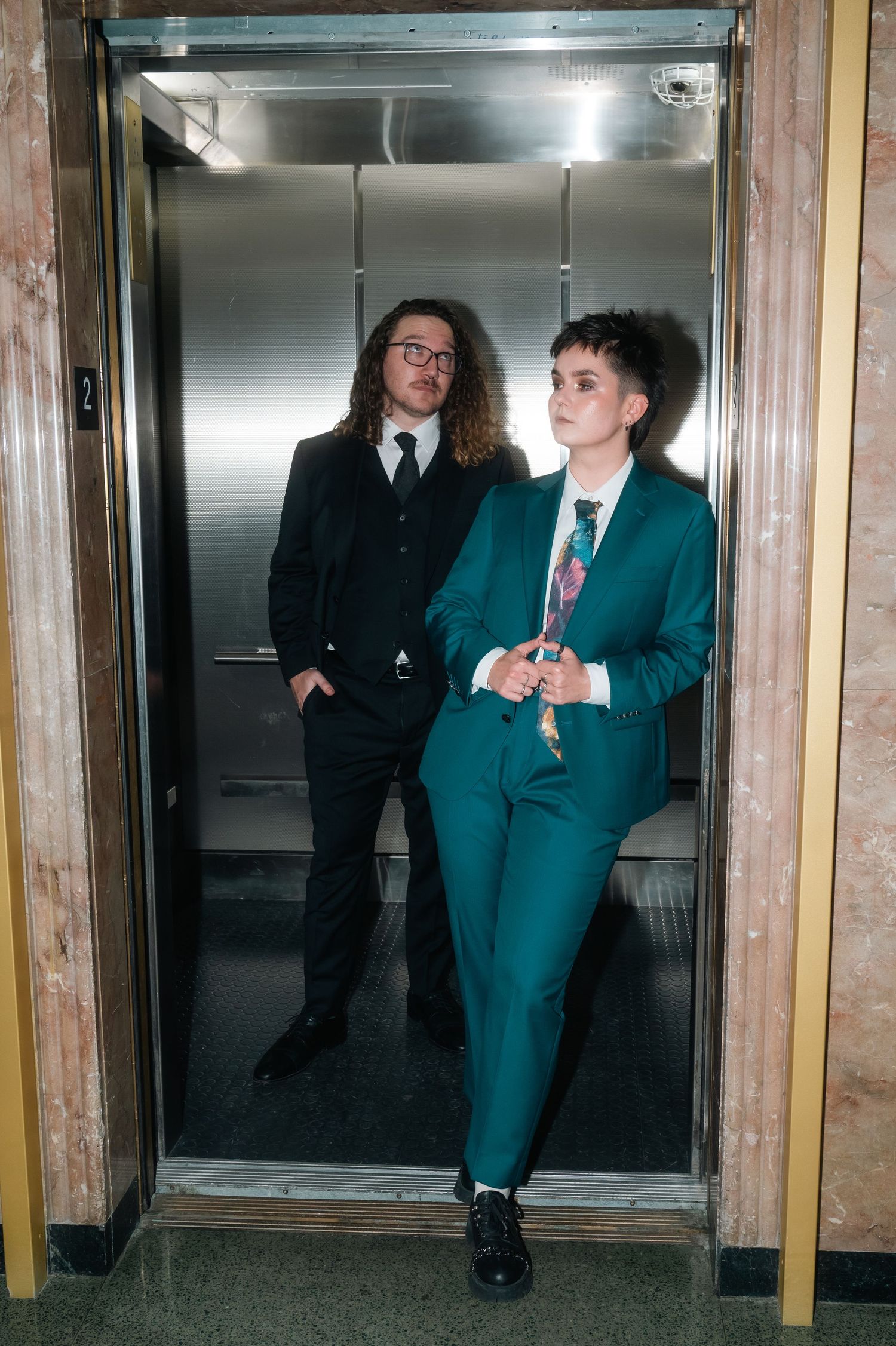 Two formally dressed individuals posing in an open elevator, one in black suit, one in teal suit with floral tie.