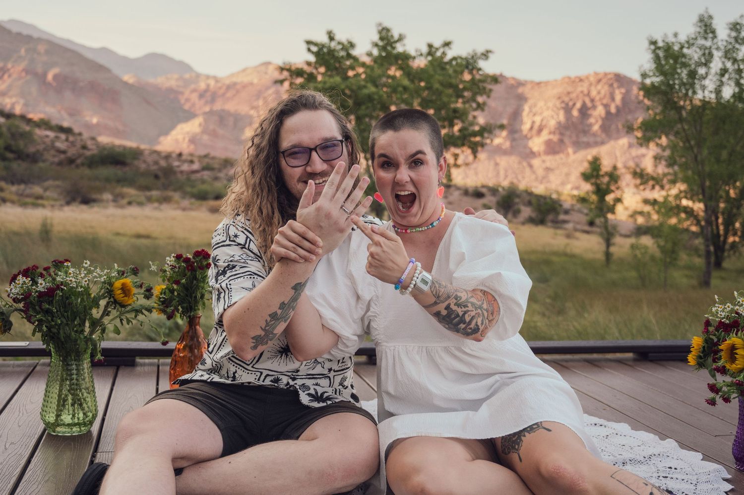 Two tattooed people joyfully showing off engagement rings on a wooden deck with mountain views at red rock canyon