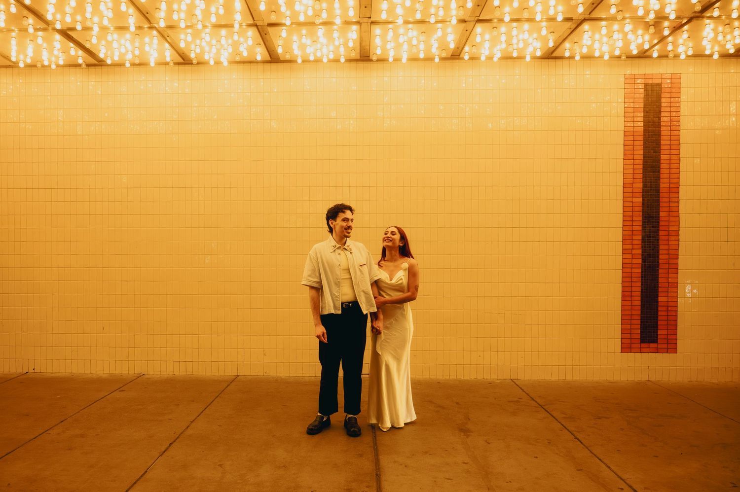 Couple smiling together in front of a yellow-lit brick wall with string lights overhead at a wedding venue.