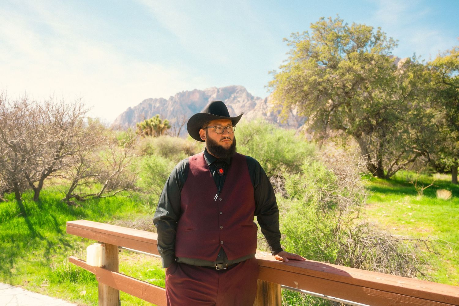Man in black cowboy hat and burgundy vest standing on wooden fence with mountain landscape background.