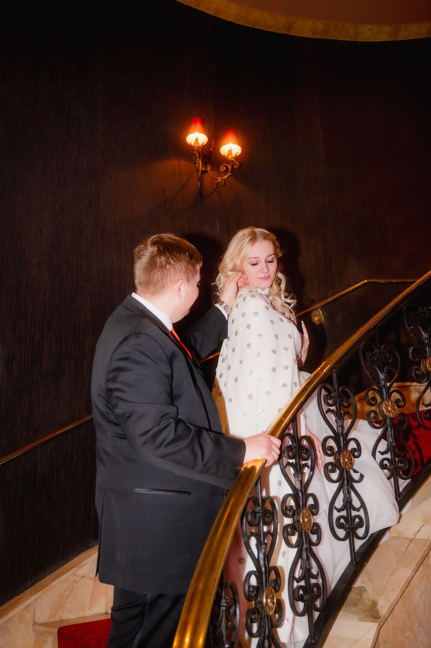 Elegant couple in formal attire on ornate staircase, bride in white gown with fur wrap, groom in black suit with red tie.