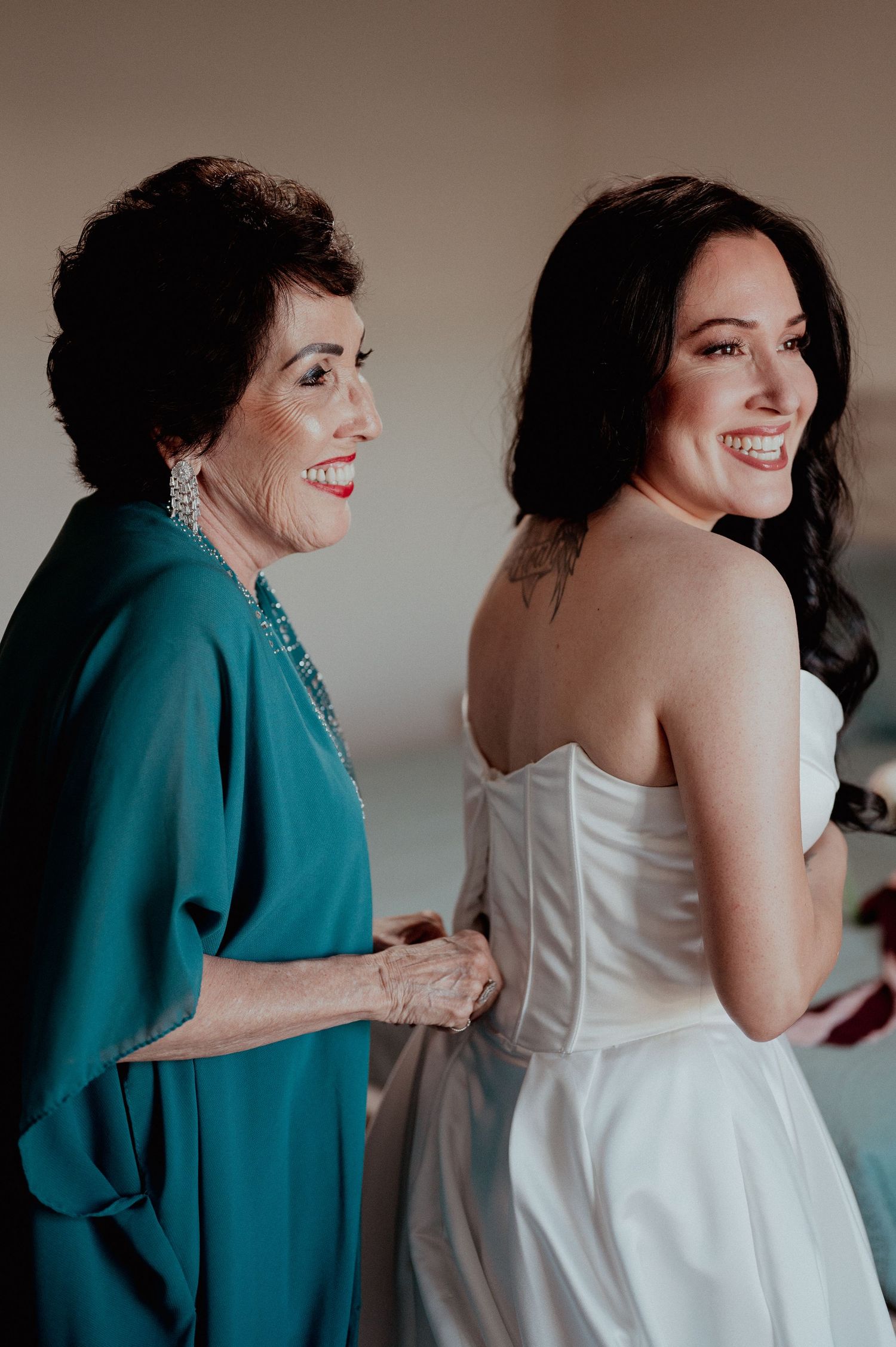 Bride in white strapless gown smiles as her mother in teal dress helps fasten her wedding dress.