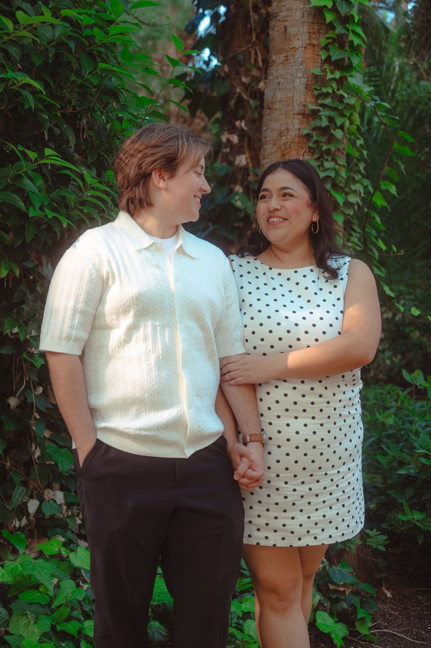 A couple holds hands and smiles at each other outdoors, surrounded by lush green ivy, dressed in elegant elopement attire