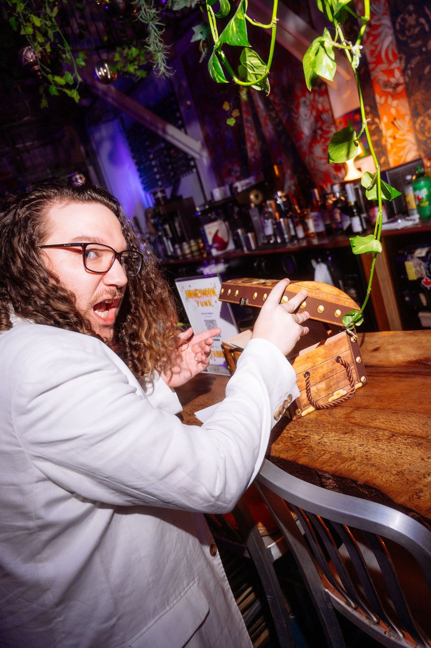 A curly-haired groom with glasses in a white jacket examines a wooden box at a bar with hanging plants.