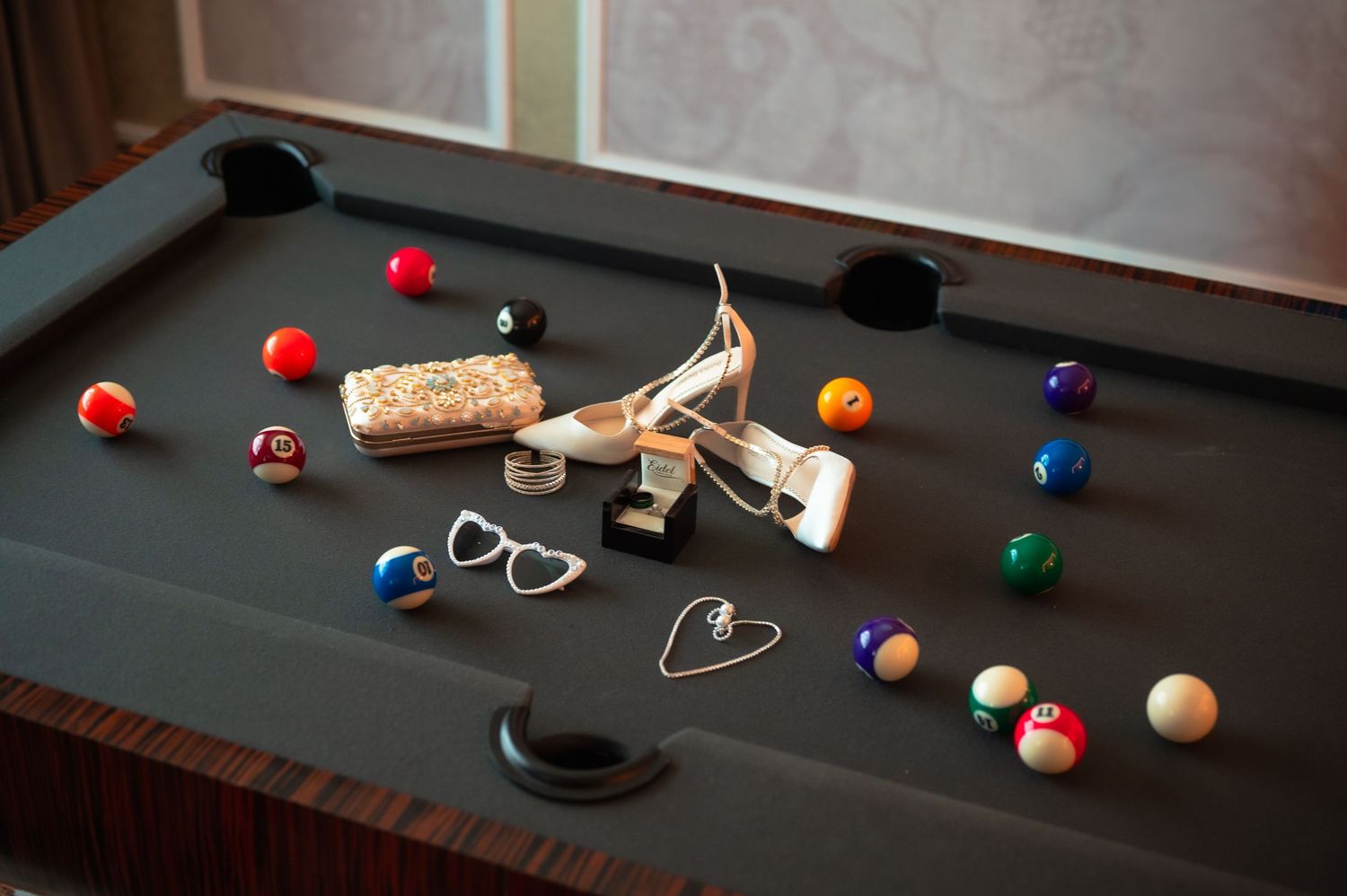 Pool table with billiard balls, high heels, heart-shaped glasses, and accessories scattered across the green felt surface.