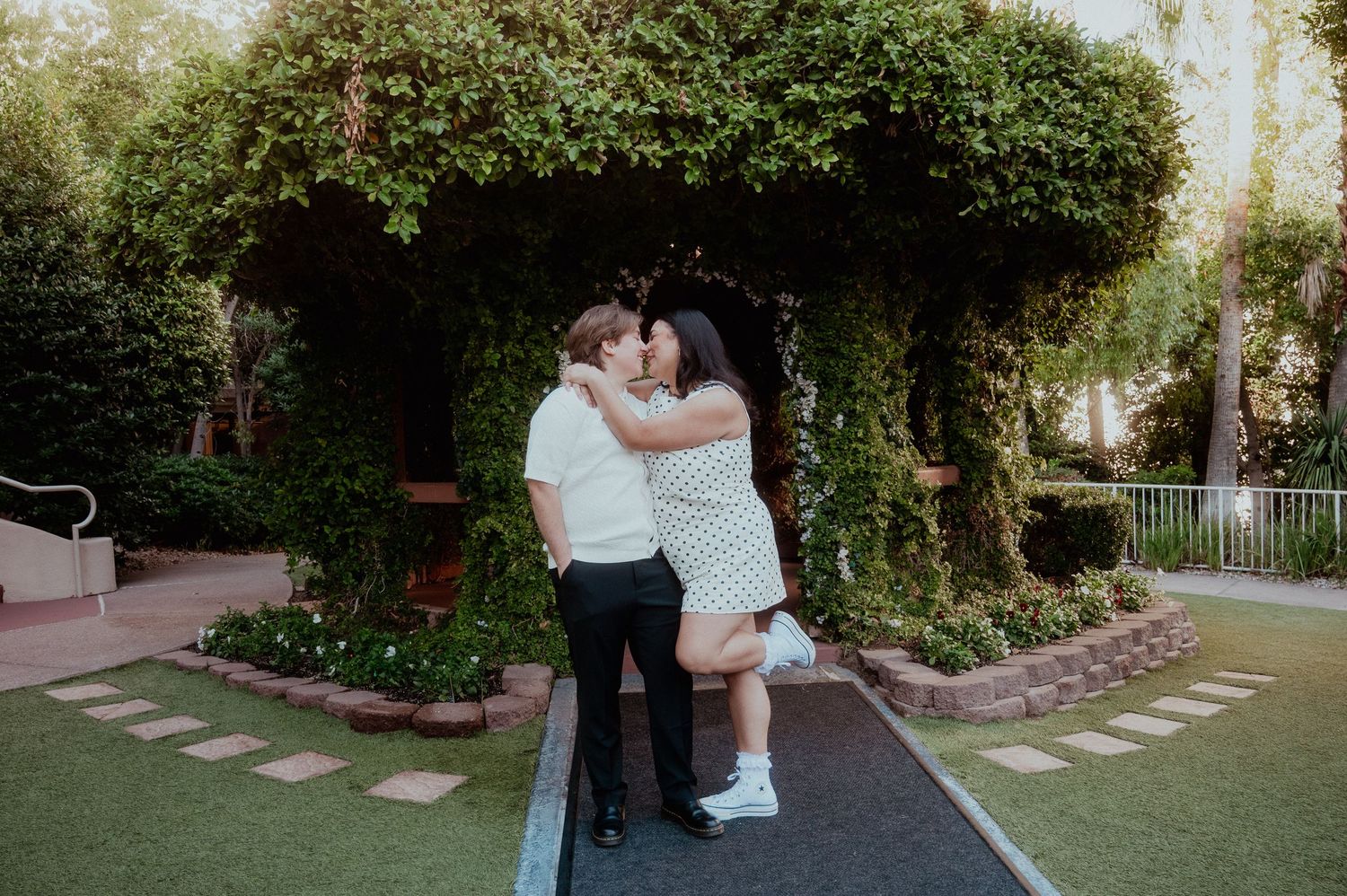 Queer Wedding Couple sharing a romantic kiss under a lush green archway on a garden pathway at sunset on the Las Vegas Strip