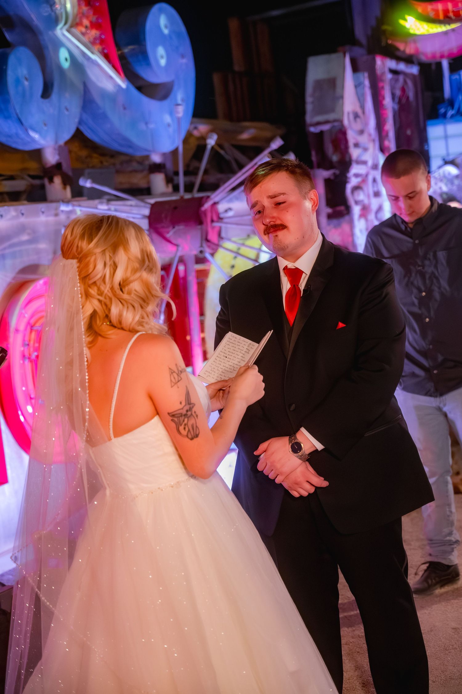 Bride reads wedding vows to emotional groom in black suit with red tie at unique neon-lit venue wedding ceremony.