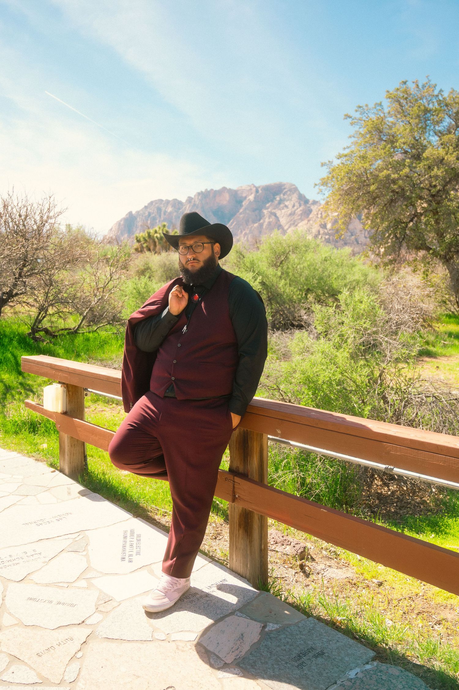 A stylish groom in a burgundy suit and black cowboy hat poses against a wooden fence with rocky mountains and greenery behind him.