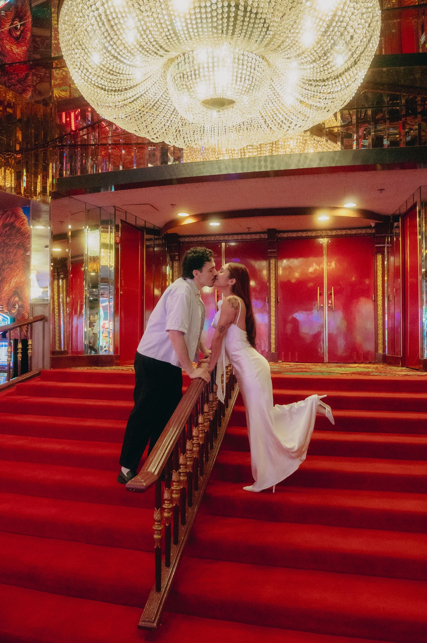 Couple sharing a romantic kiss on red carpeted stairs under a grand crystal chandelier in an elegant venue.