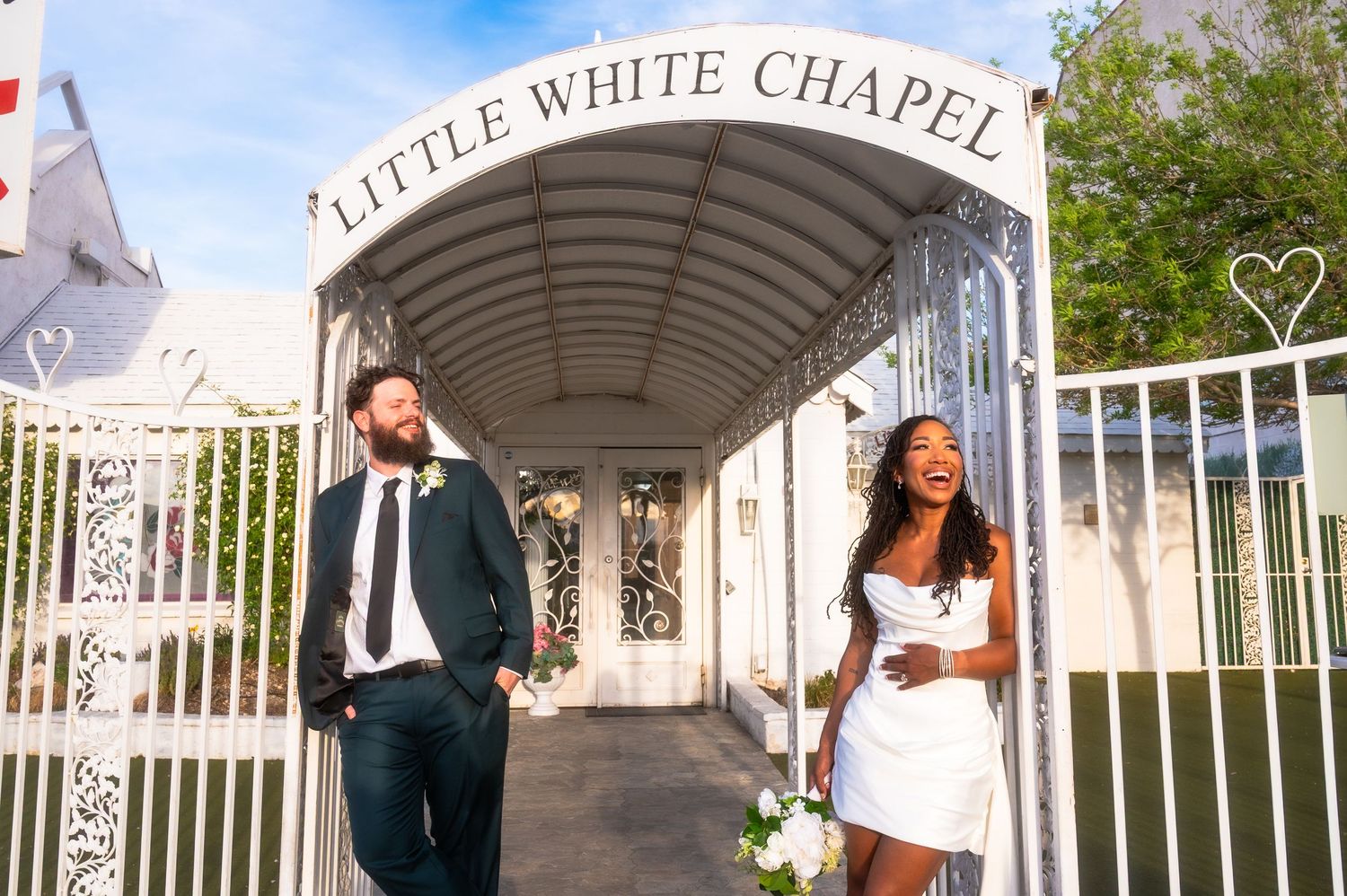 Couple laughing outside Little White Chapel in Las Vegas, bride in white mini dress holding bouquet, groom in dark suit.