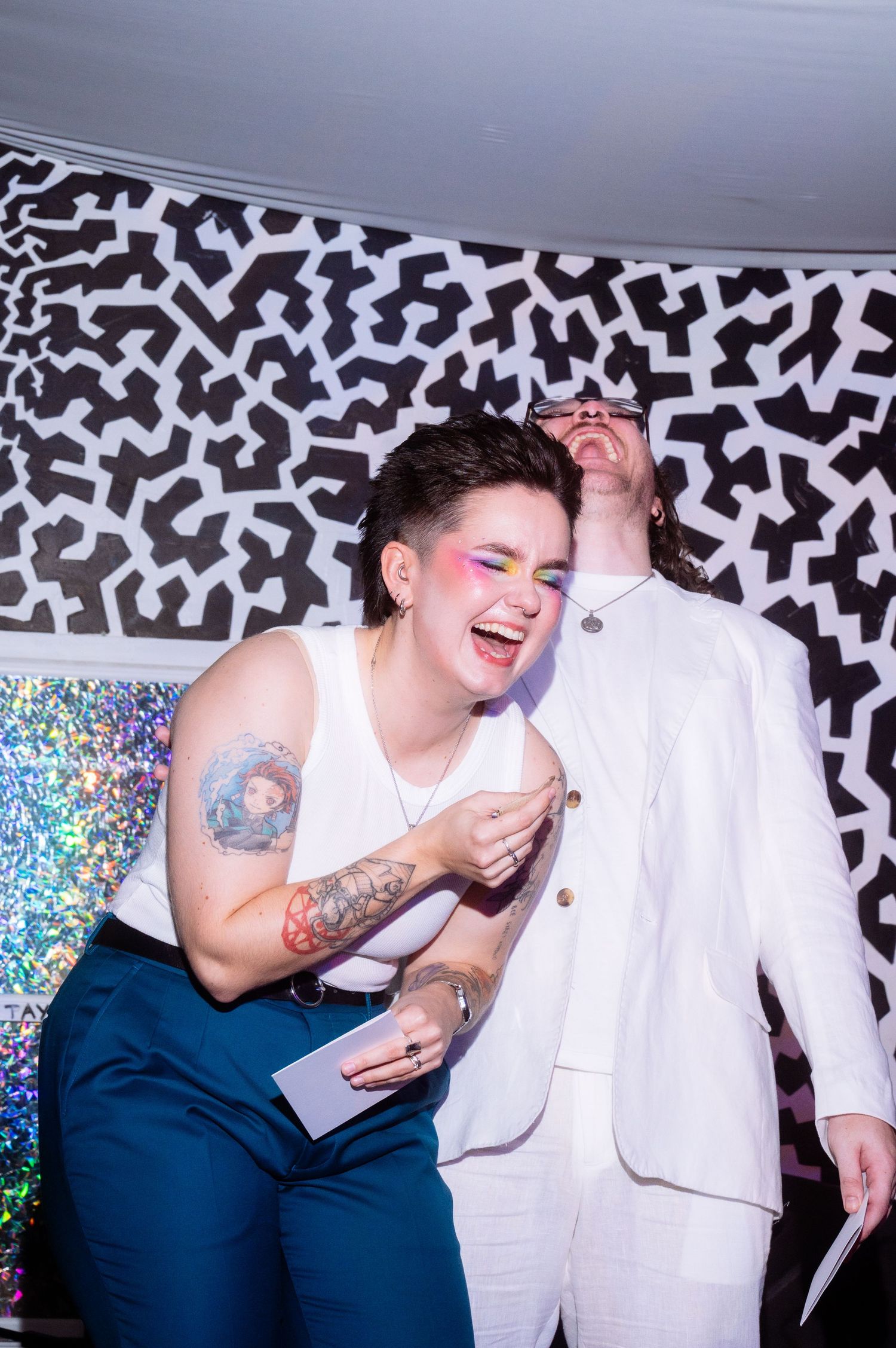 Two people laughing at a party, one with colorful makeup and tattoos, posing in front of a bold black and white patterned backdrop.