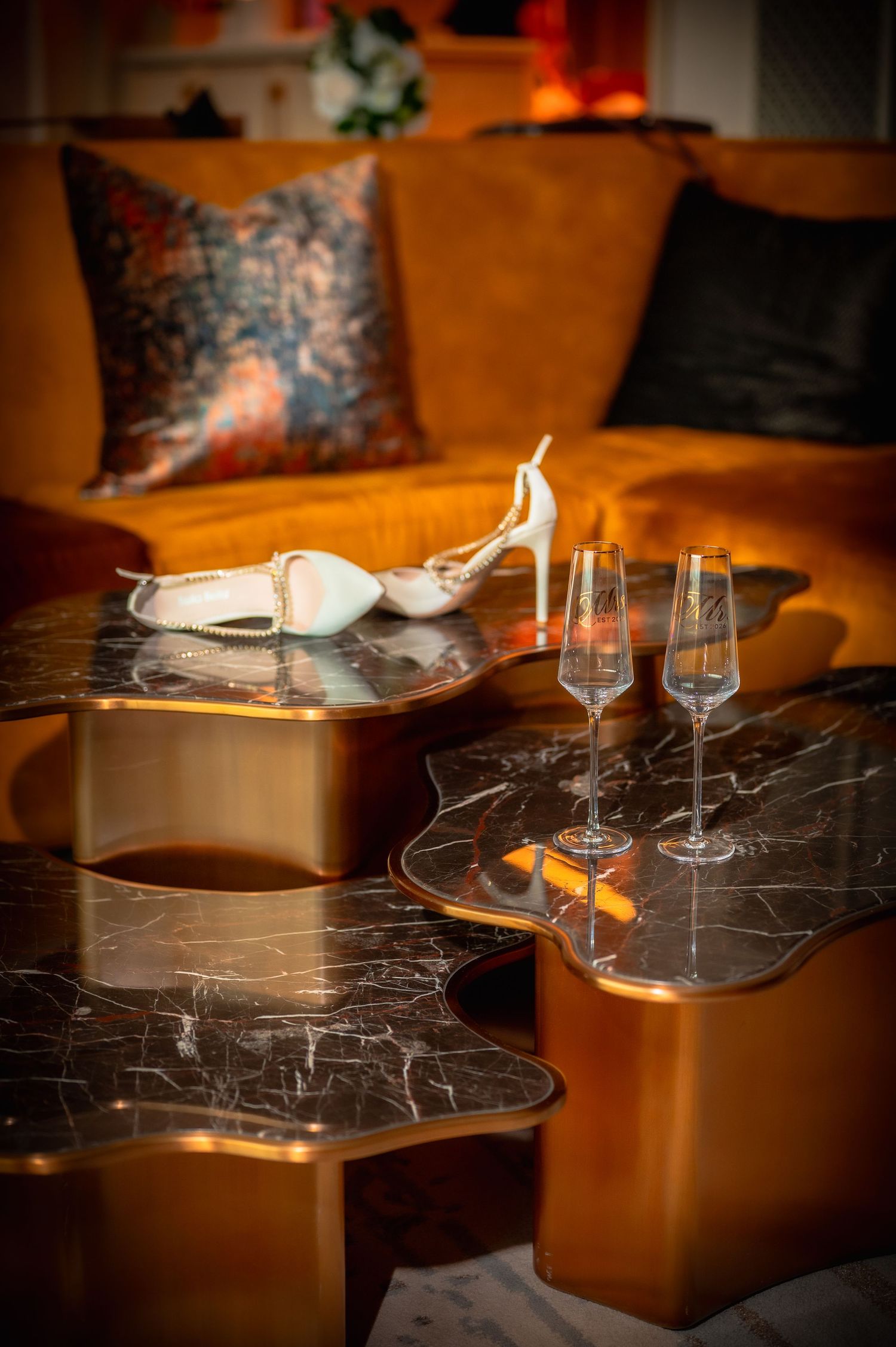 Elegant marble coffee table with champagne flutes and white heels on gold base beside an orange velvet sofa.