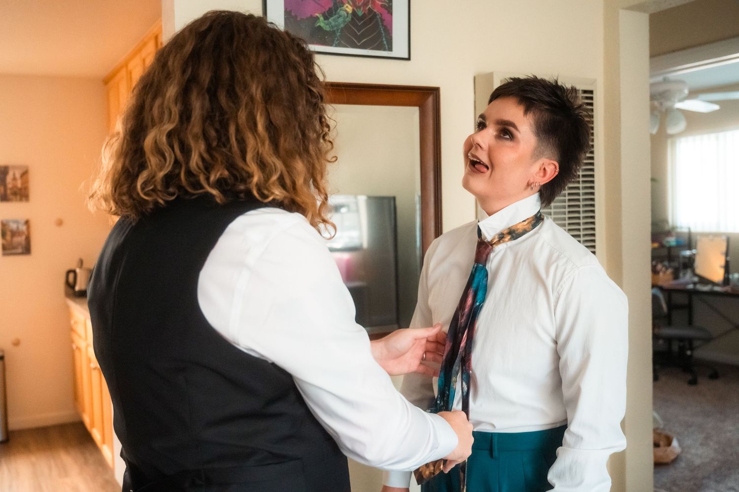 Two people getting ready in formal wear, one helping the other tie a necktie before their wedding.