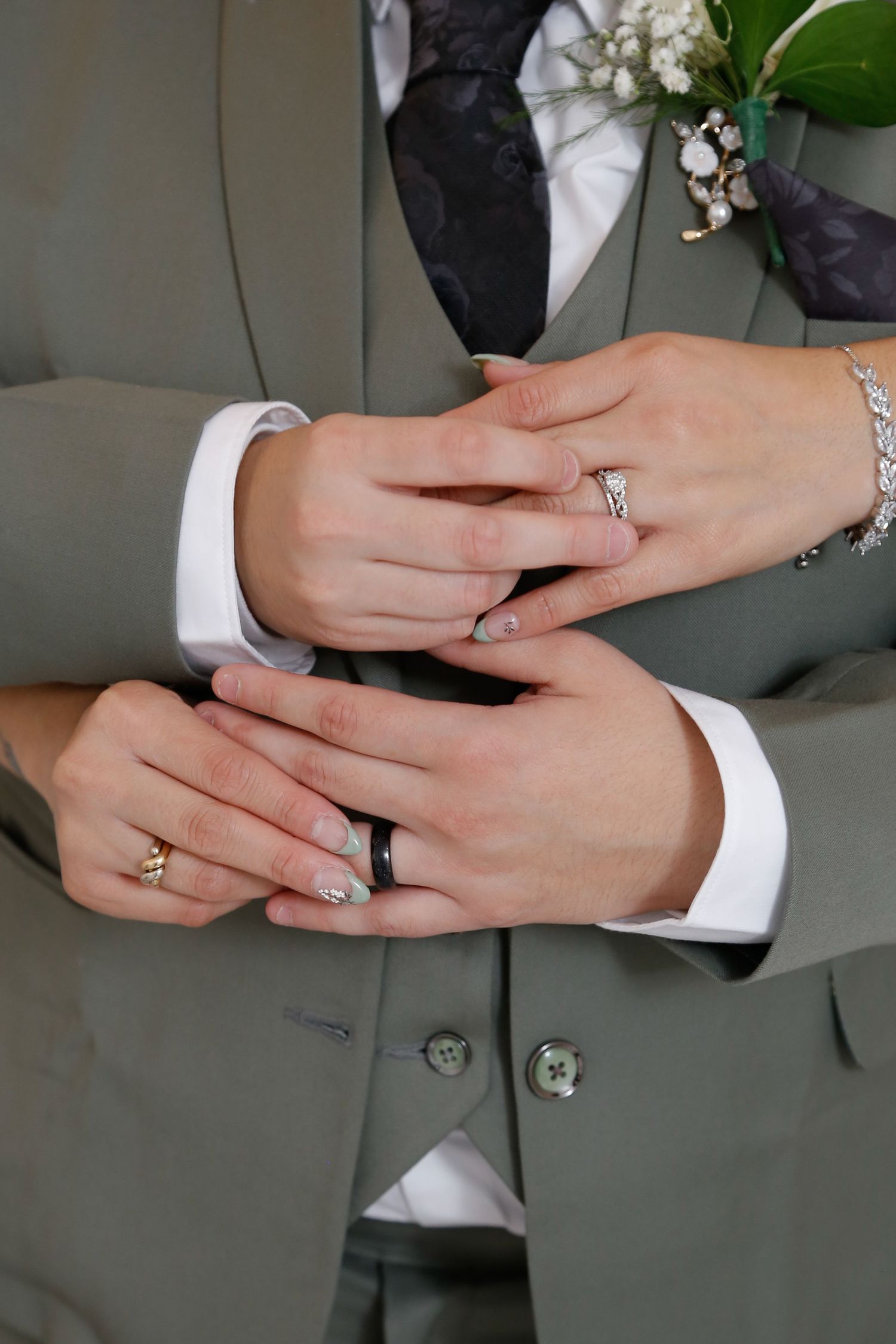 Newlywed couple displaying wedding rings, groom in gray suit with boutonniere, bride wearing diamond ring and bracelet.