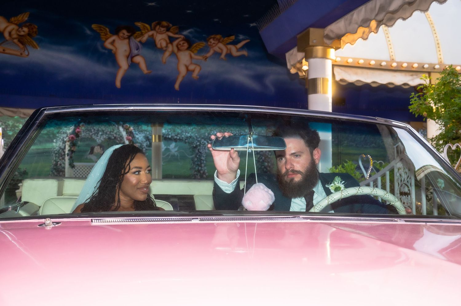 Newlywed couple in a pink car at a Las Vegas wedding chapel with cherub mural backdrop.