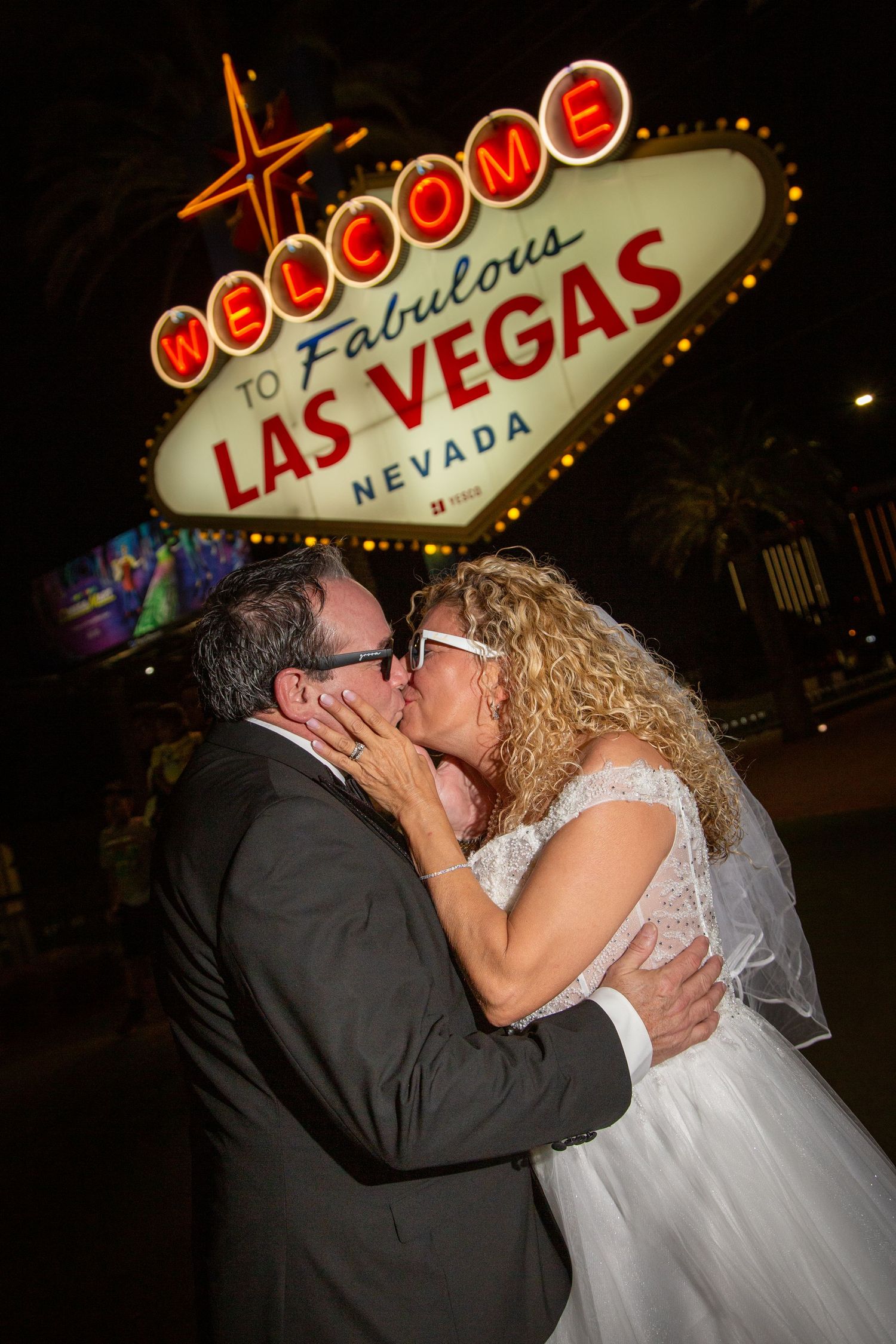 Newlywed couple sharing a kiss in front of the iconic Welcome to Fabulous Las Vegas Nevada sign at night.