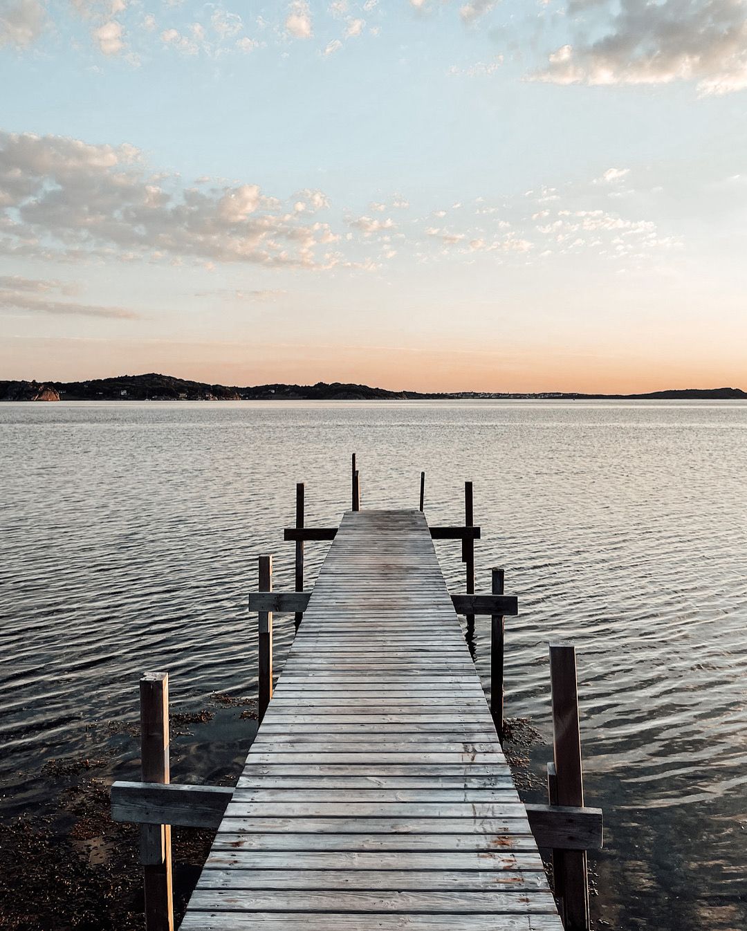 Wooden dock extending into calm lake water at sunset with pastel sky and distant shoreline.