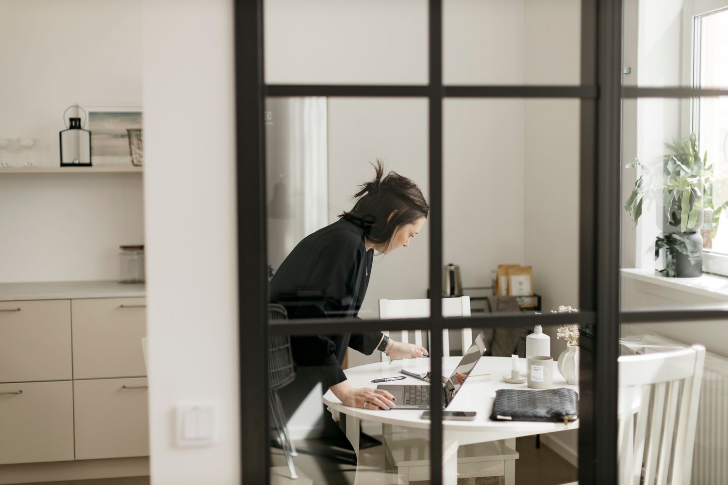 Woman in black outfit leaning over table working on laptop in modern Scandinavian home office.