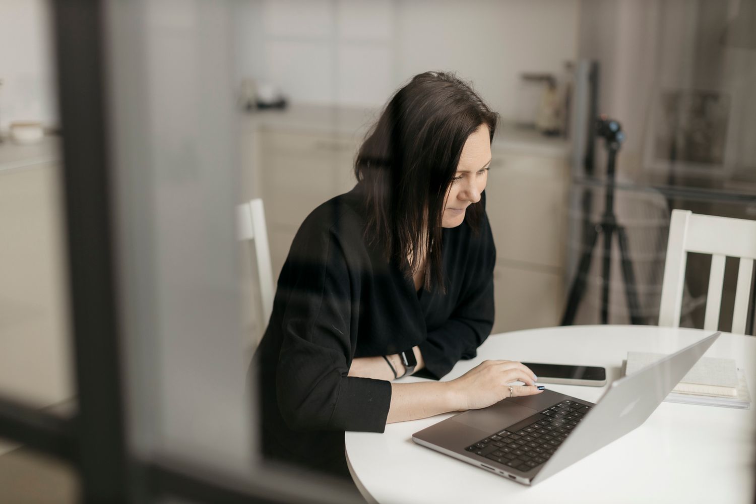 Dark-haired businesswoman in black blazer working on laptop at round white table by window.
