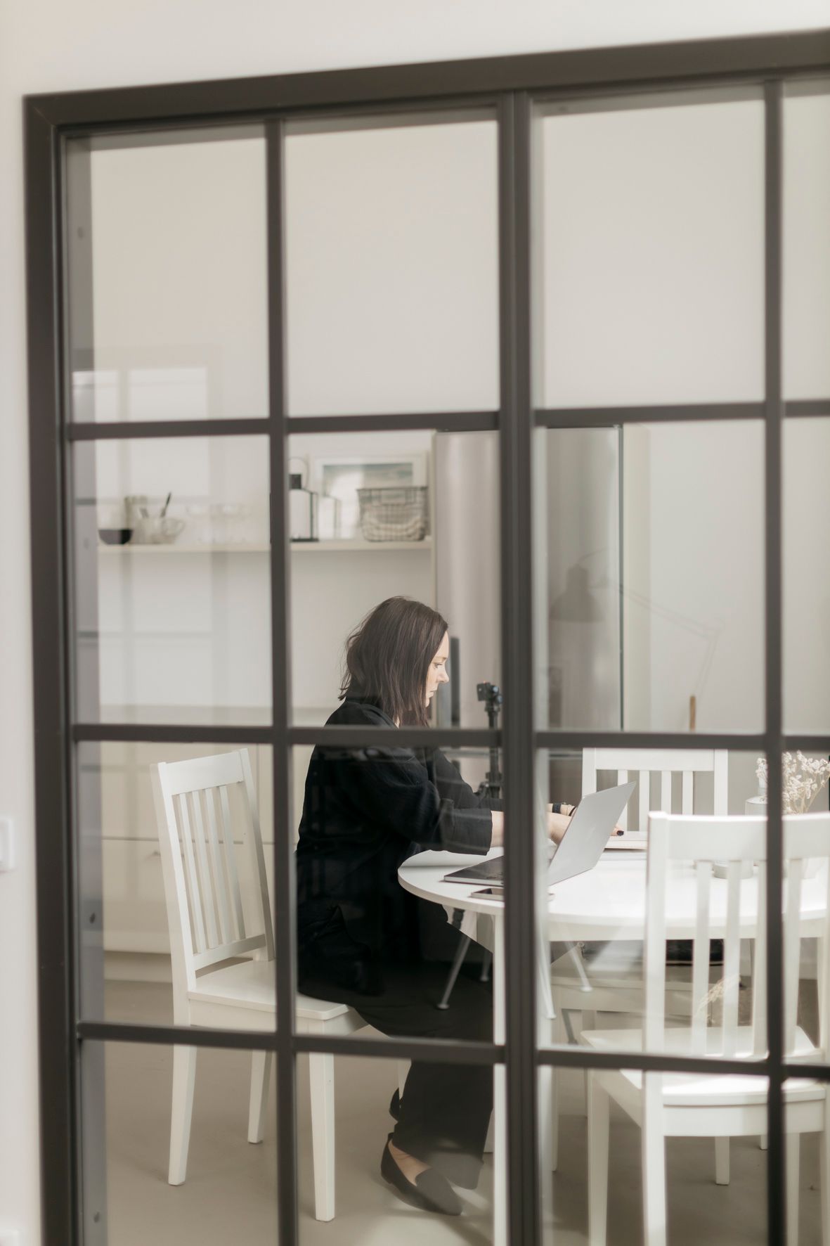 Woman working on laptop at white dining table viewed through black-framed glass partition doors.
