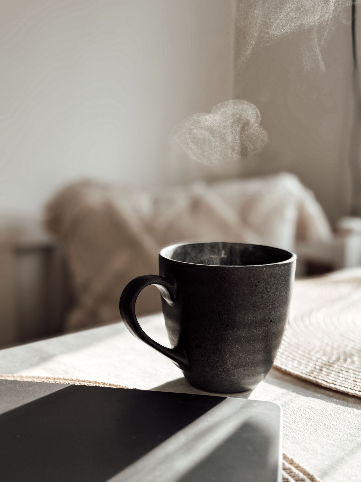 A steaming black ceramic mug on a table beside a laptop, with a cozy blurred background.