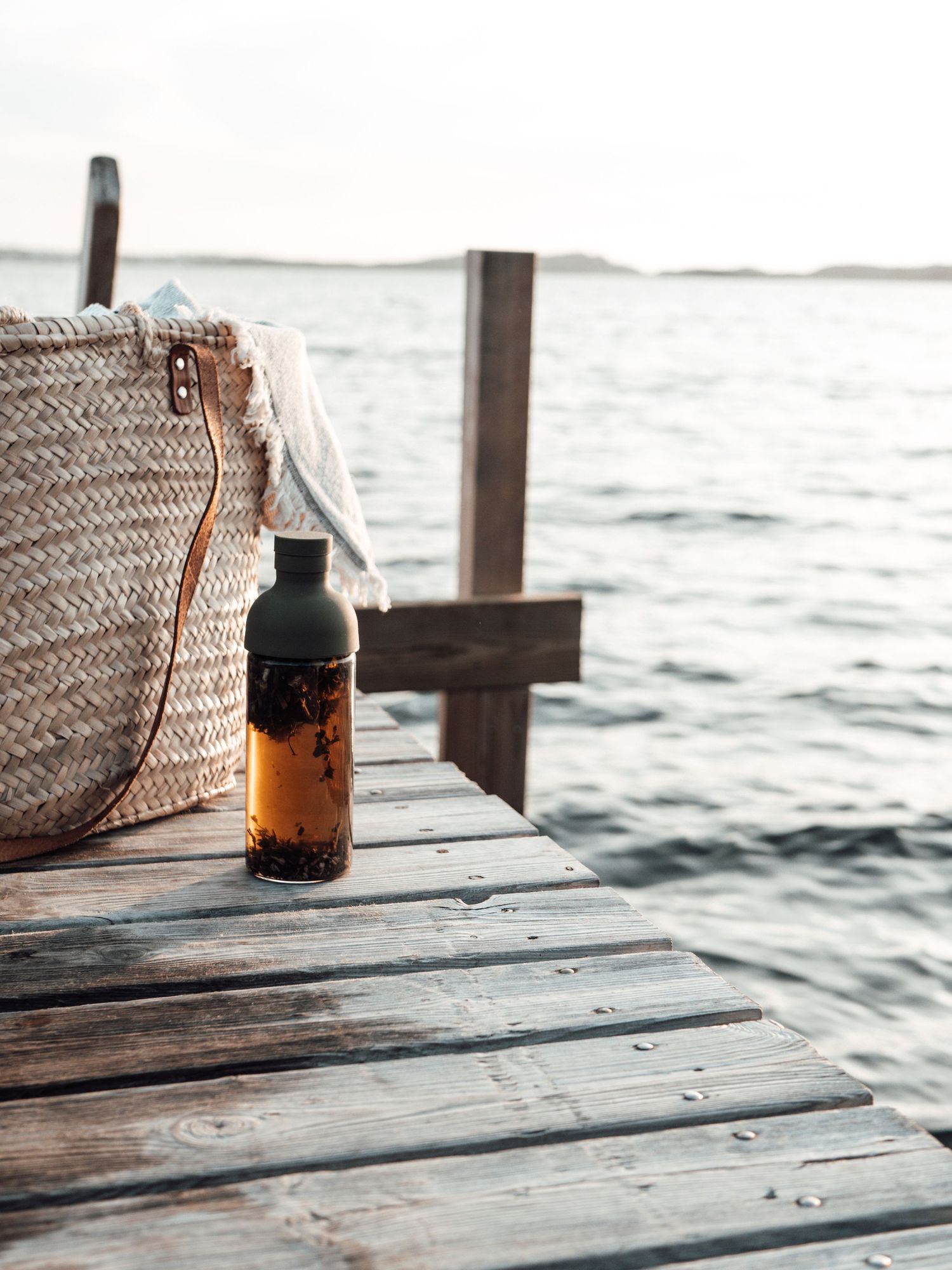 Straw beach bag and dark water bottle on wooden dock by calm sea at sunset.