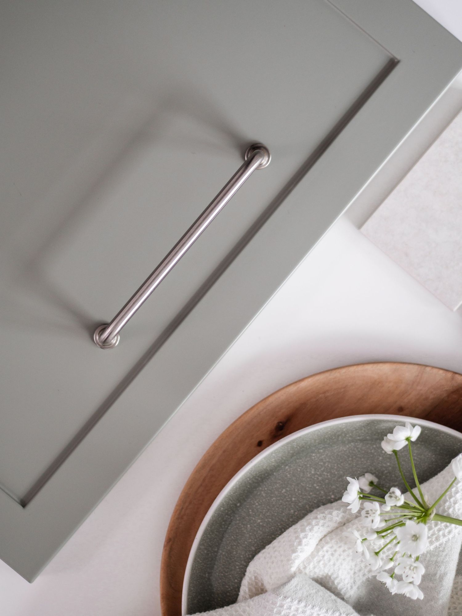 Close-up of a sage green kitchen cabinet drawer with a brushed nickel bar handle and ceramic bowl with white flowers.