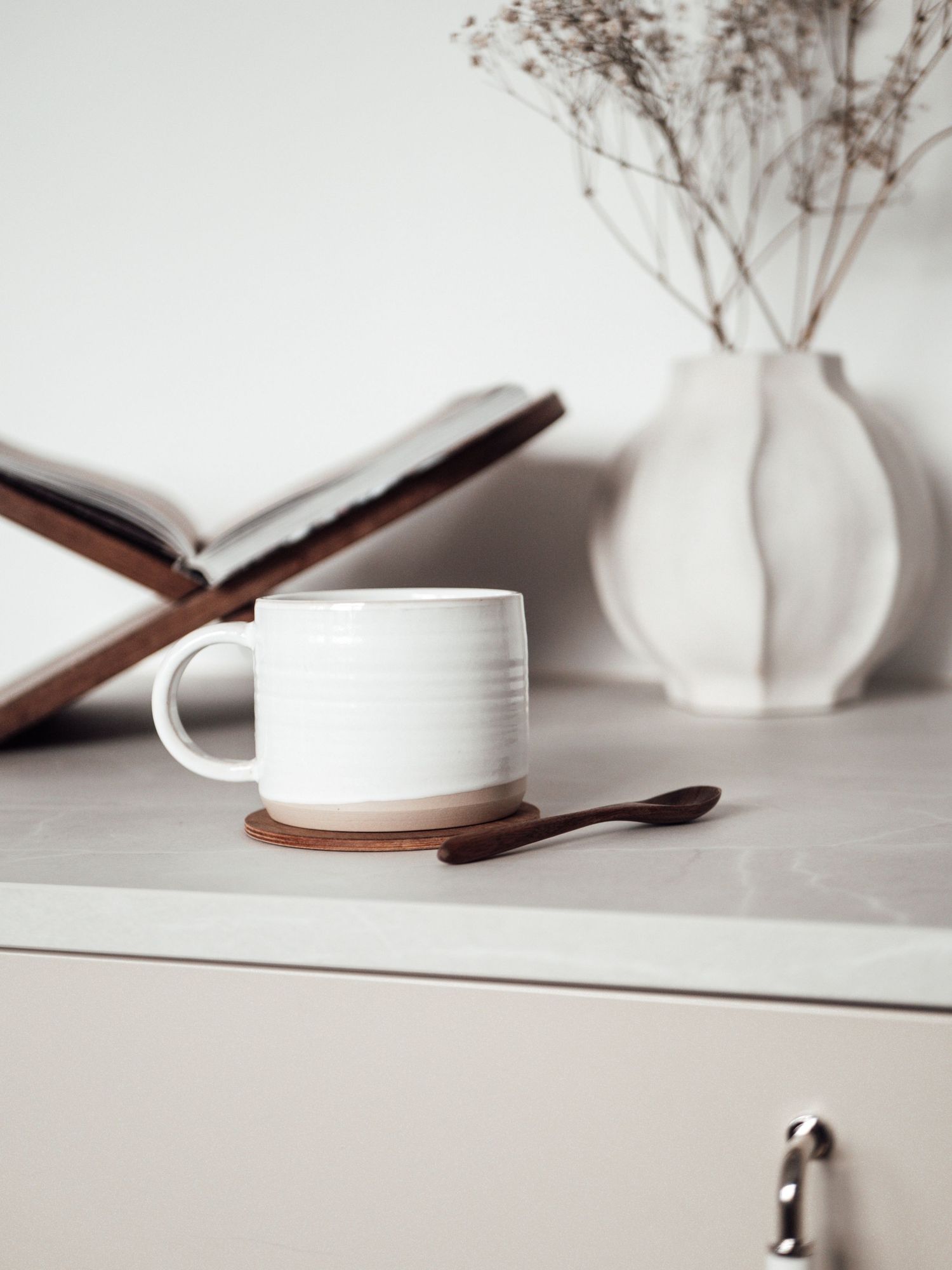 White ribbed ceramic mug on wooden coaster beside open book and white vase with dried flowers on dresser.
