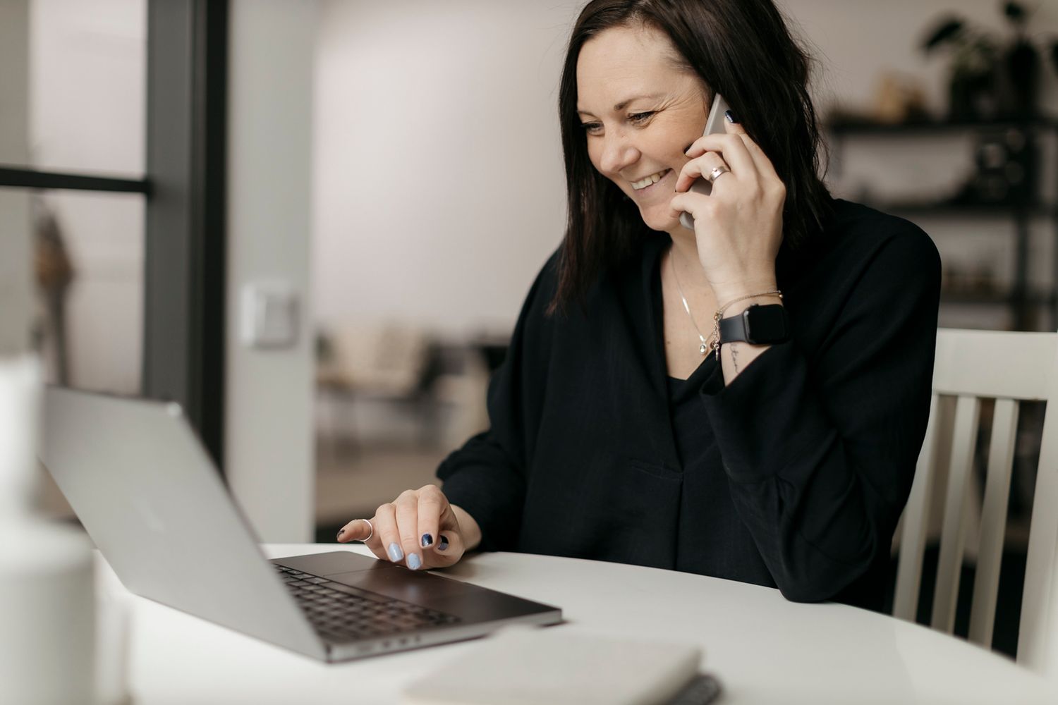 Smiling businesswoman in black blazer talking on phone while using laptop at white table.