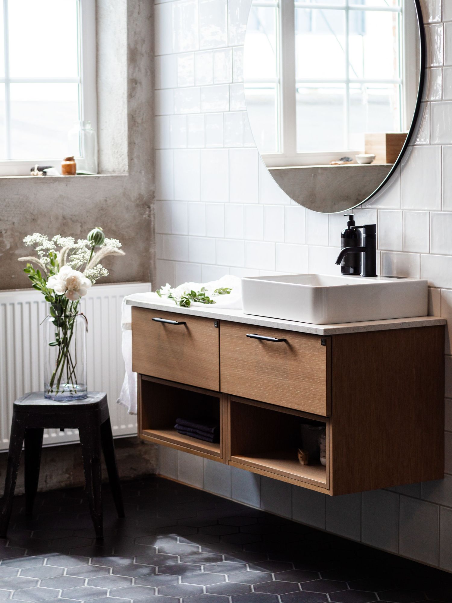 Modern Scandinavian bathroom with wooden floating vanity, round mirror, white vessel sink, and fresh flowers.