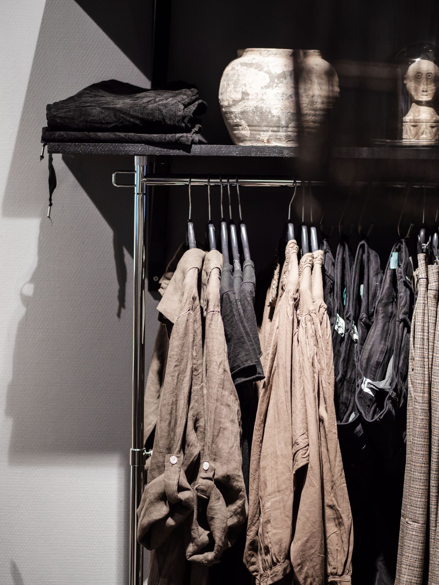 Dark retail clothing rack with neutral linen garments hanging on hangers beside rustic ceramic vases.