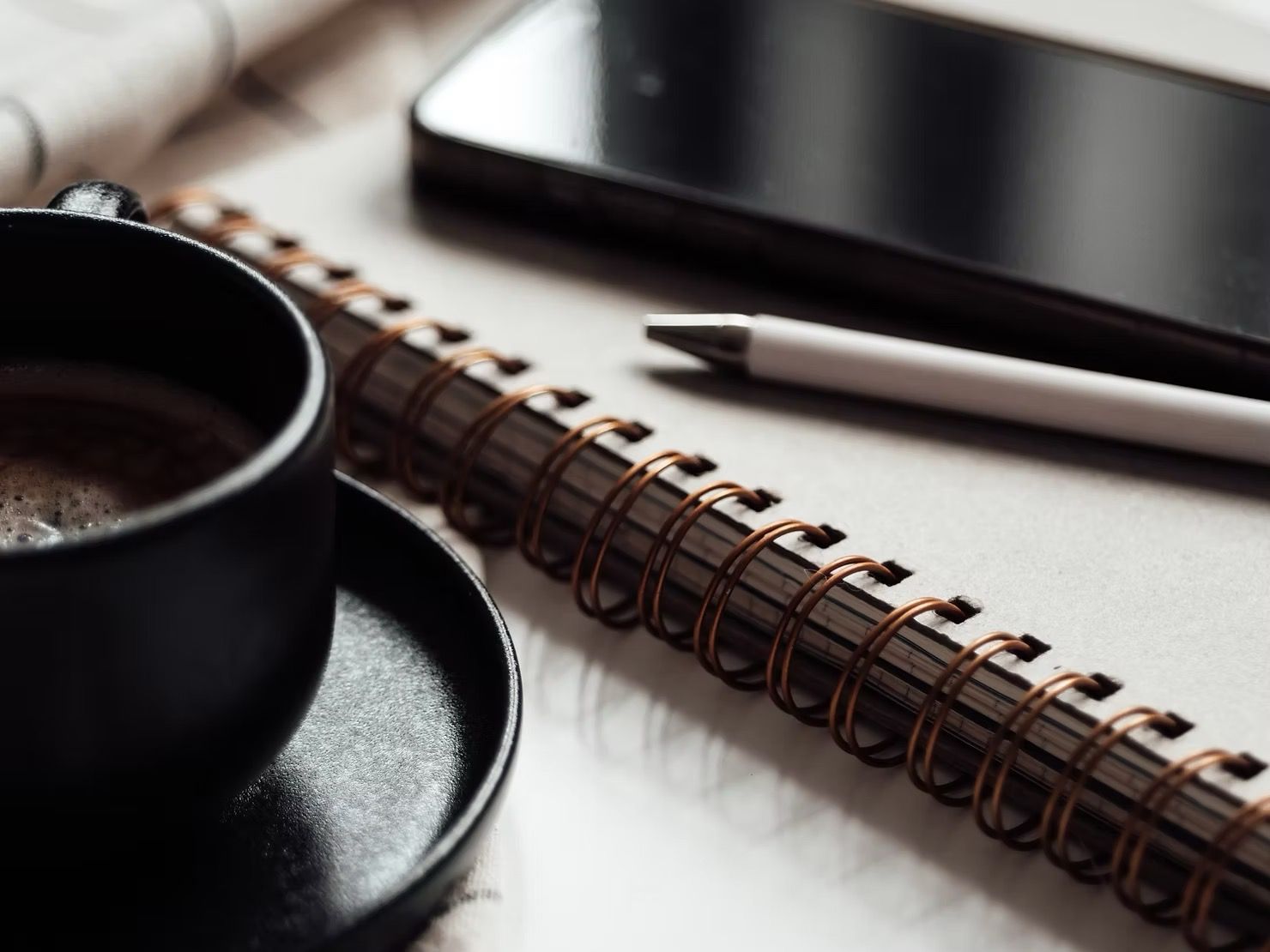 Black coffee cup, spiral notebook, white pen and smartphone on textured white surface.