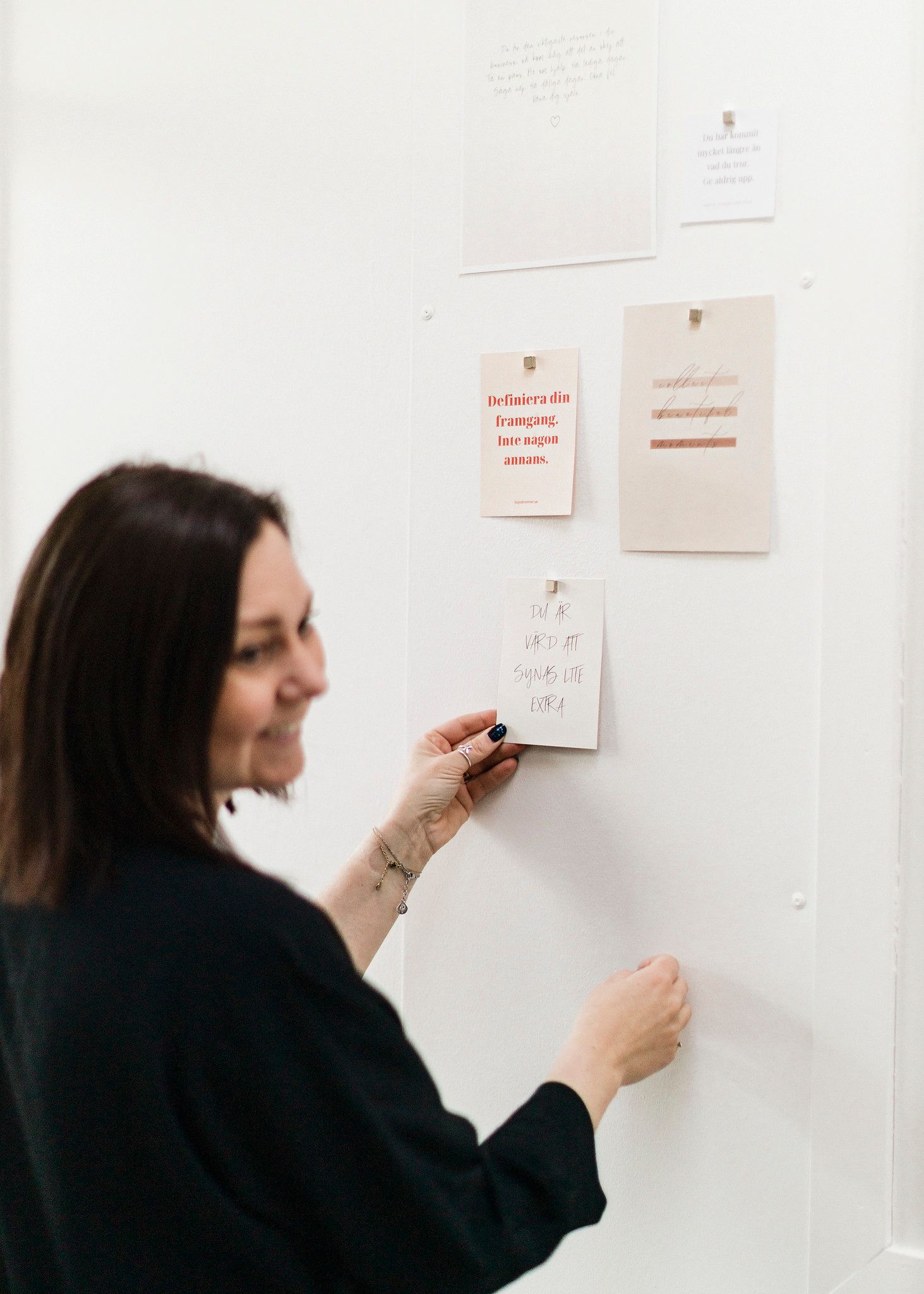 Woman in black outfit pinning notes and cards to a white mood board wall.
