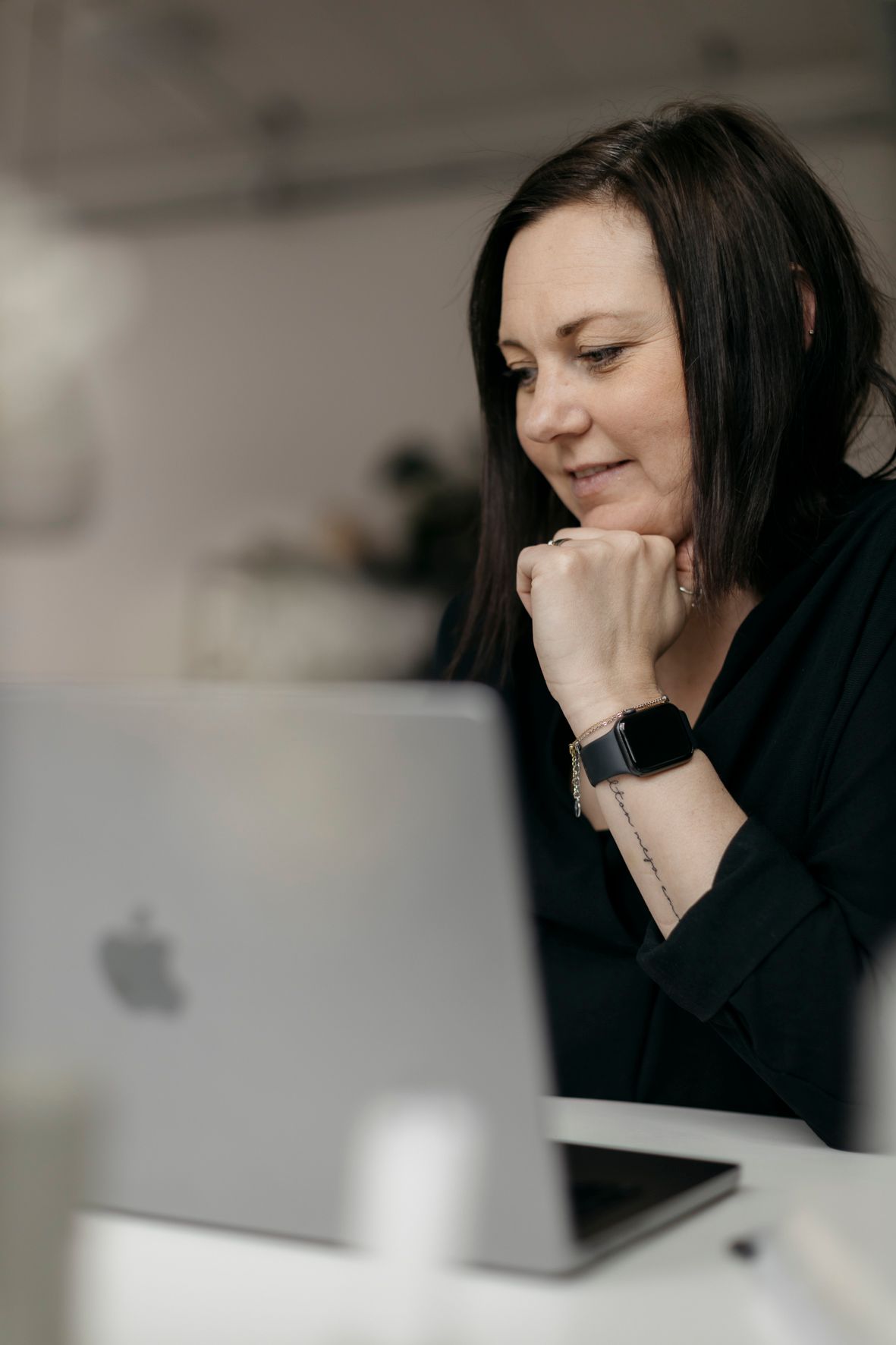 Dark-haired professional woman in black top working focused on Apple MacBook laptop at desk.