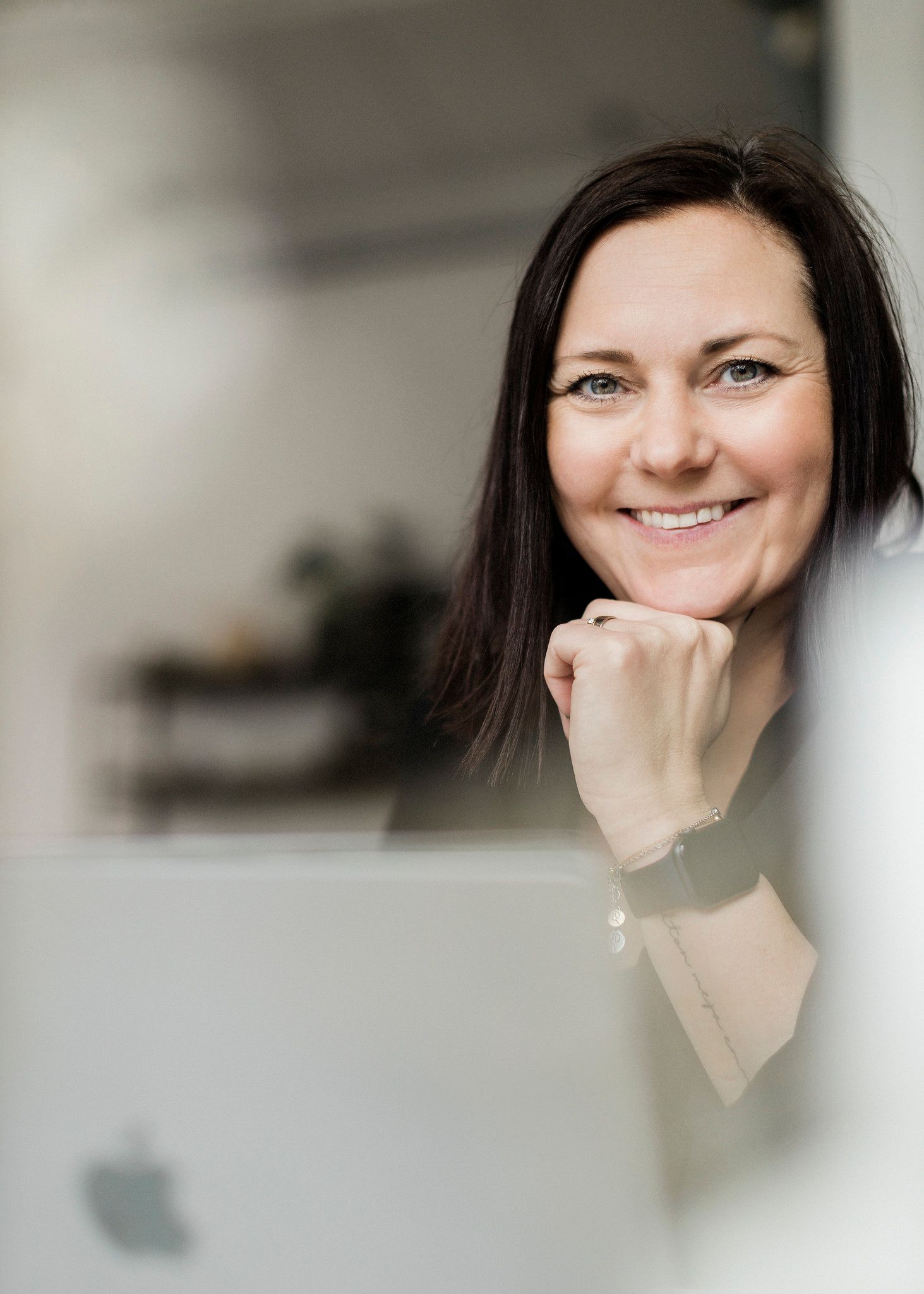 Smiling dark-haired woman resting chin on hand working at Apple MacBook laptop in bright office.