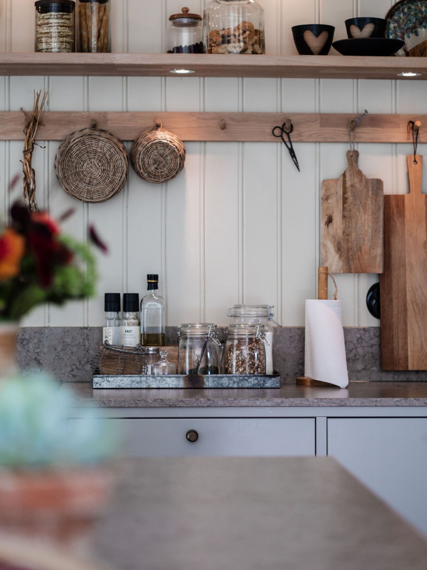 Rustic kitchen counter with glass jars, spices, wooden boards, and wicker wall decor.