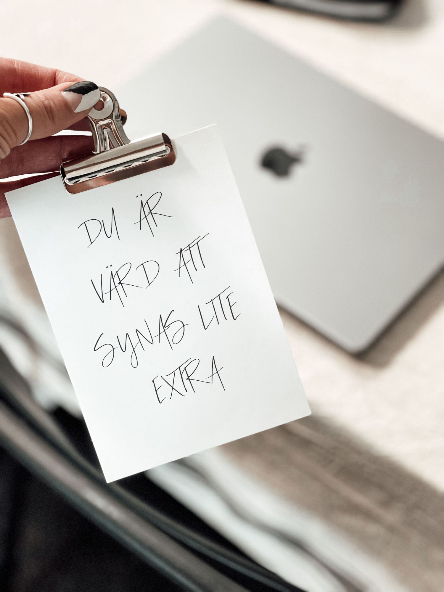 Handwritten Swedish note reading 'Du ar vard att synas lite extra' held with binder clip near MacBook.