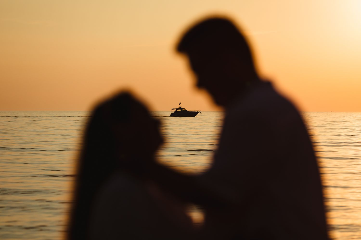 Silhouetted figures against golden sunset over ocean with boat in distance.