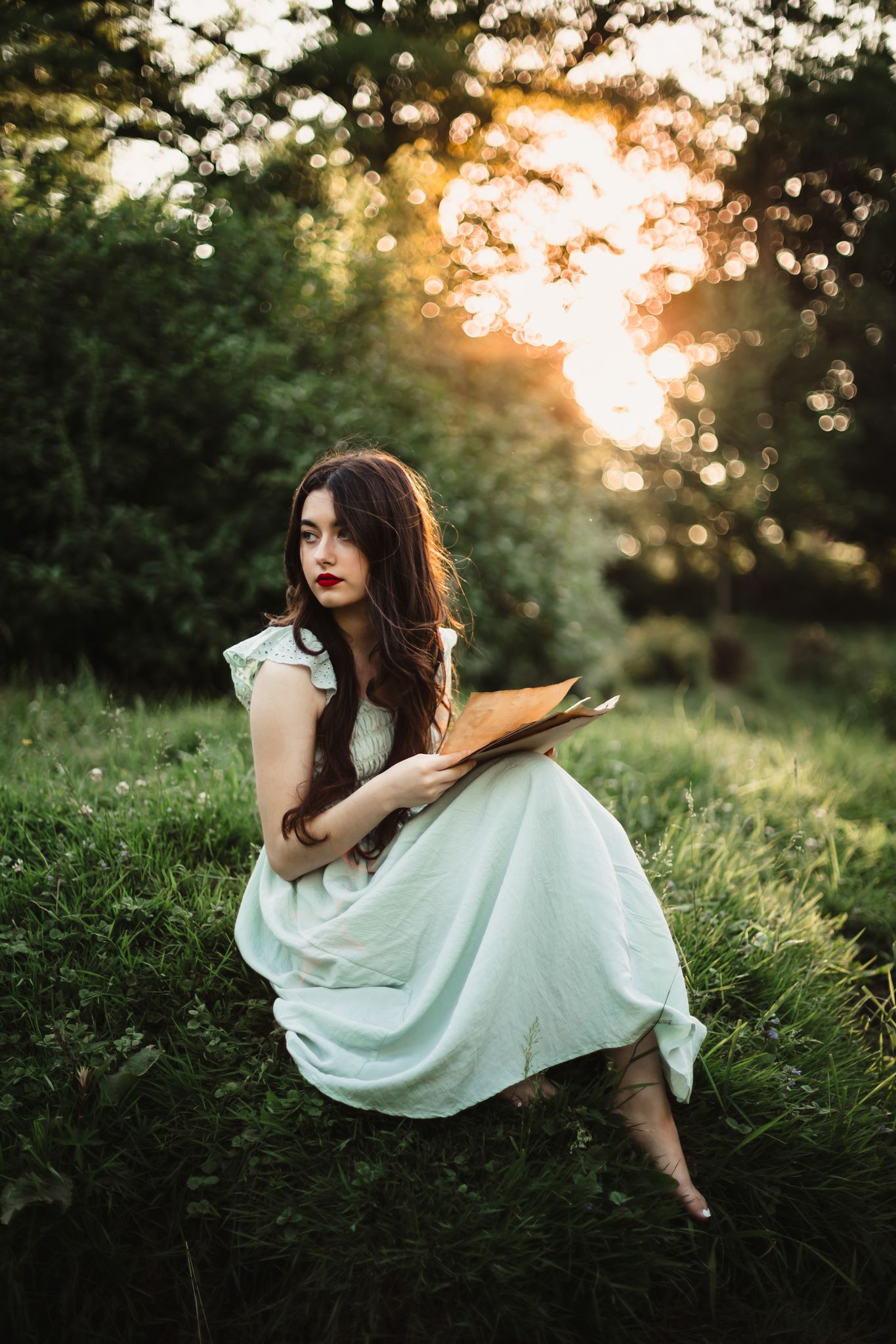 Figure in flowing white dress seated on grass during golden sunset.
