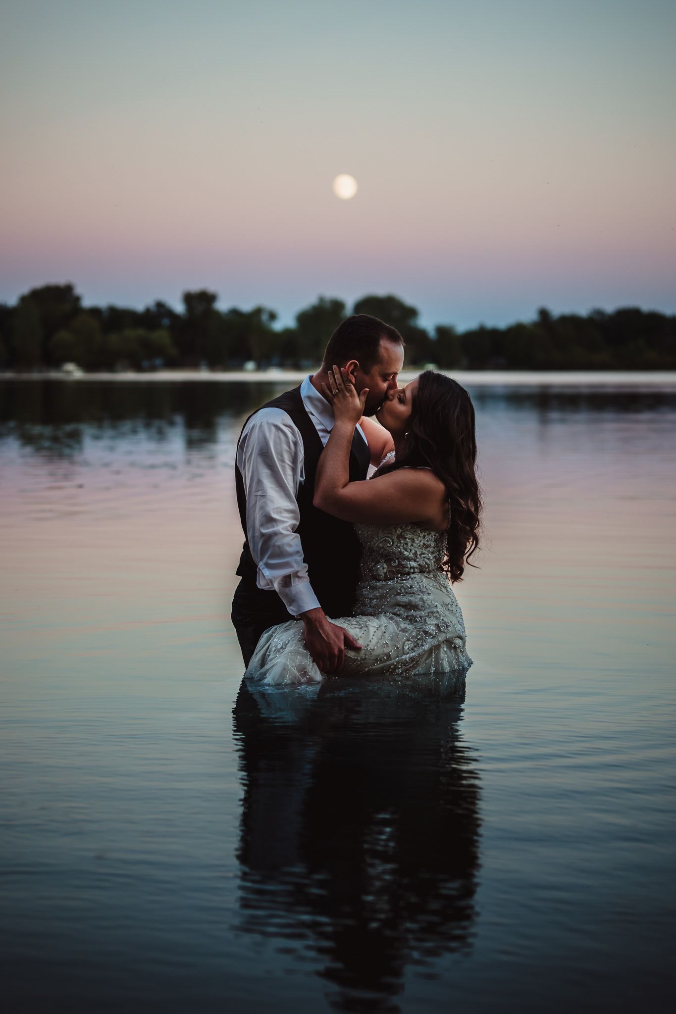 A couple shares a romantic kiss while standing in calm lake waters at sunset.
