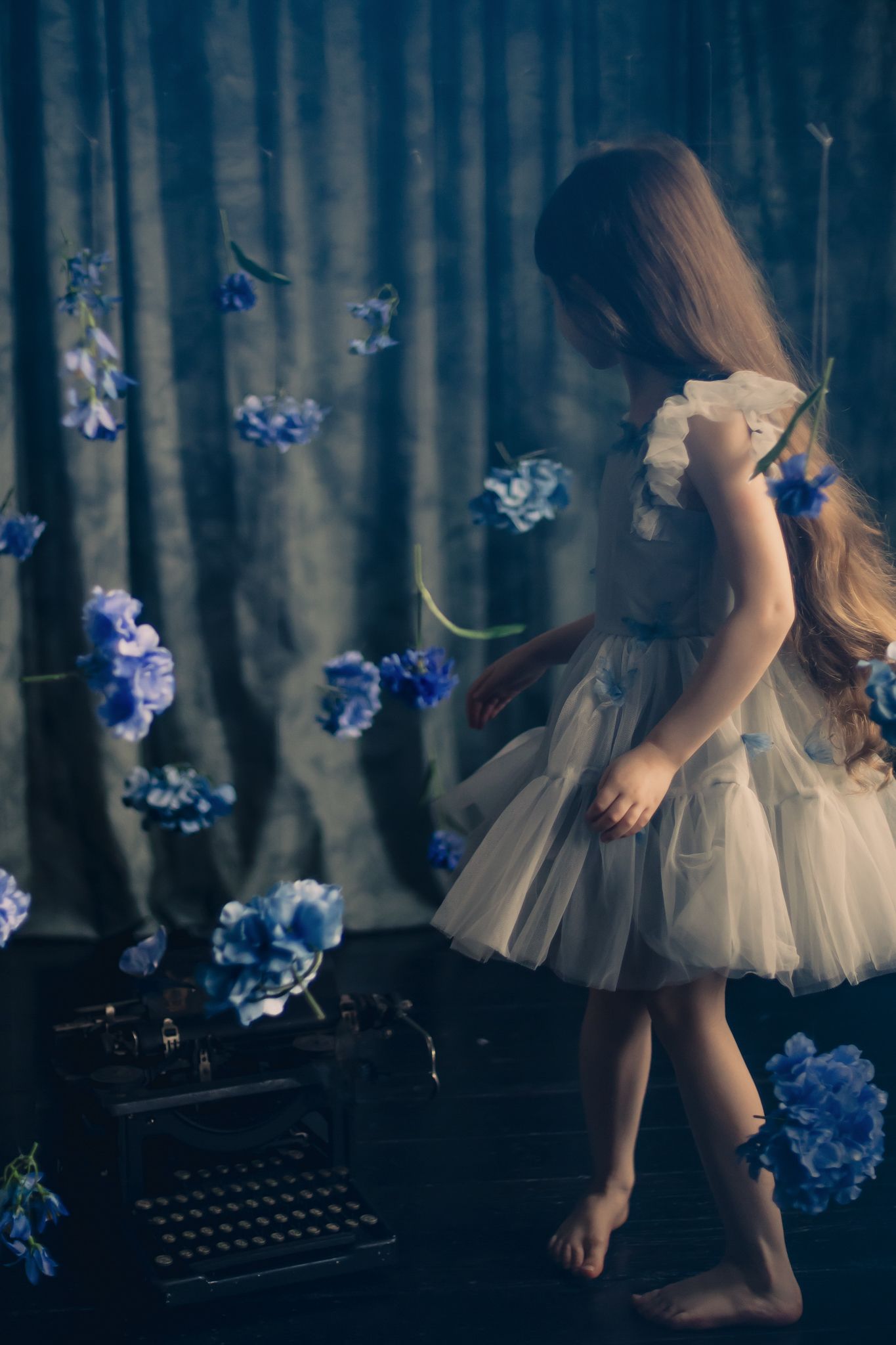 A dreamy scene with floating blue flowers, vintage typewriter, and a figure in a white tulle dress dancing barefoot.