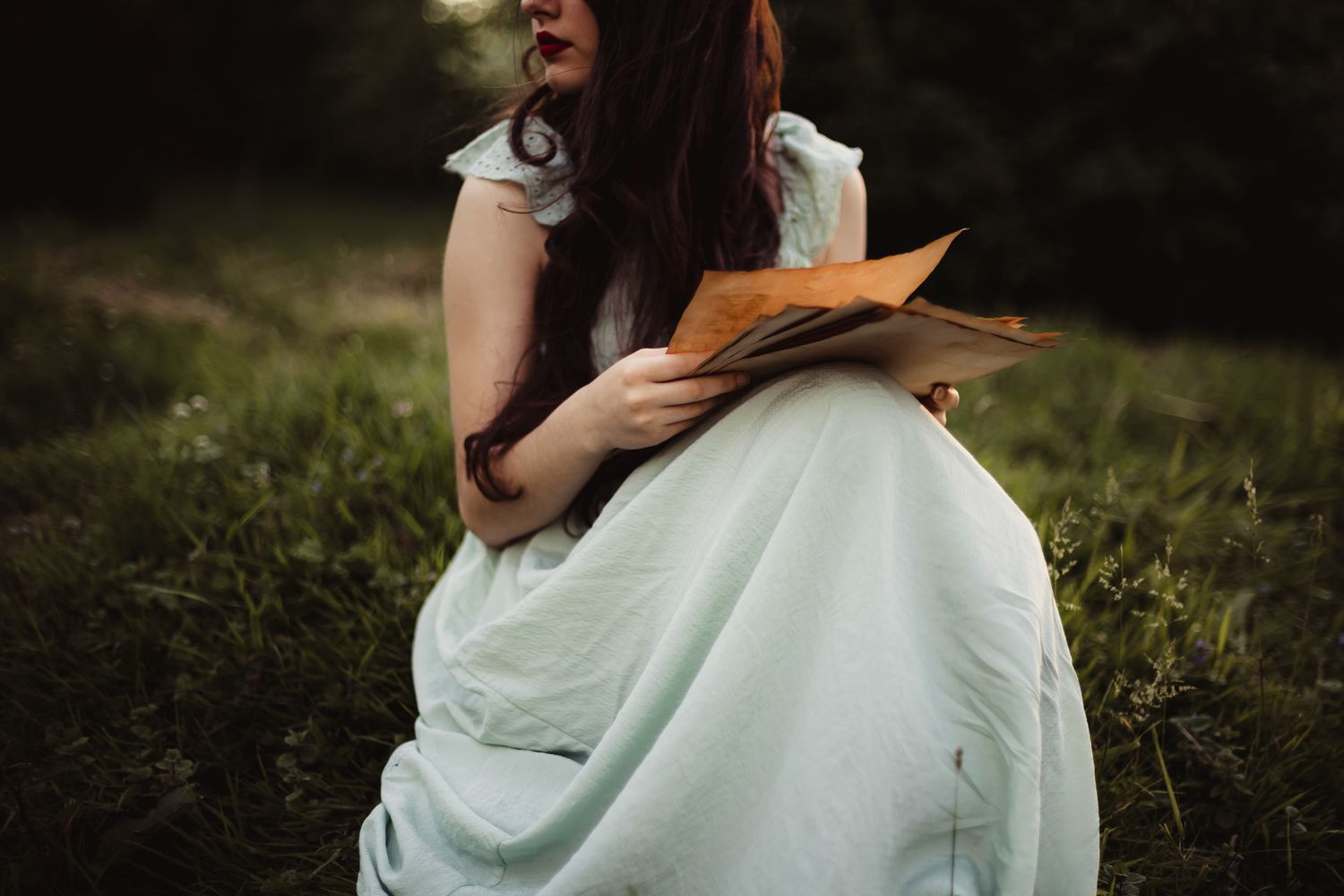 A person in a flowing mint green dress sits gracefully in a sunlit meadow with long grass.