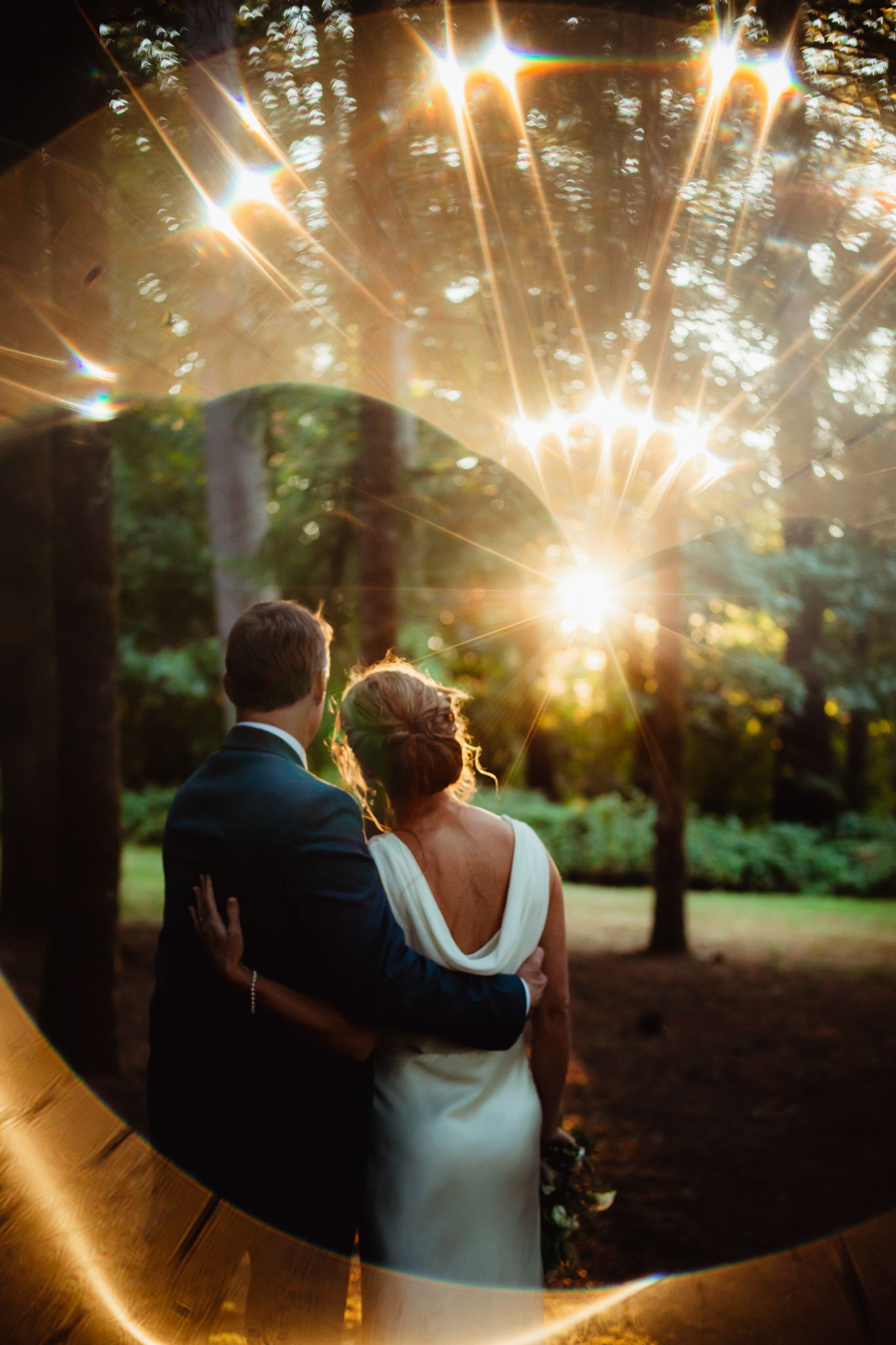 A couple stands together in a wooded area at sunset with dramatic lens flares creating a romantic atmosphere.