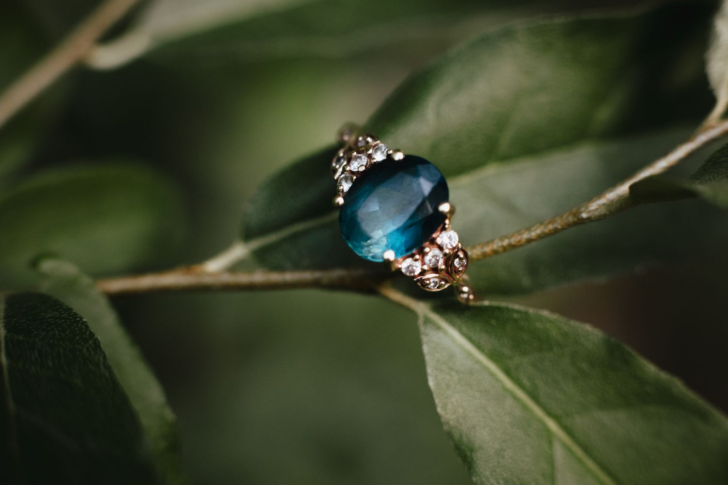 Vintage style engagement ring with blue gemstone and diamond accents rests on green leaves in natural light.