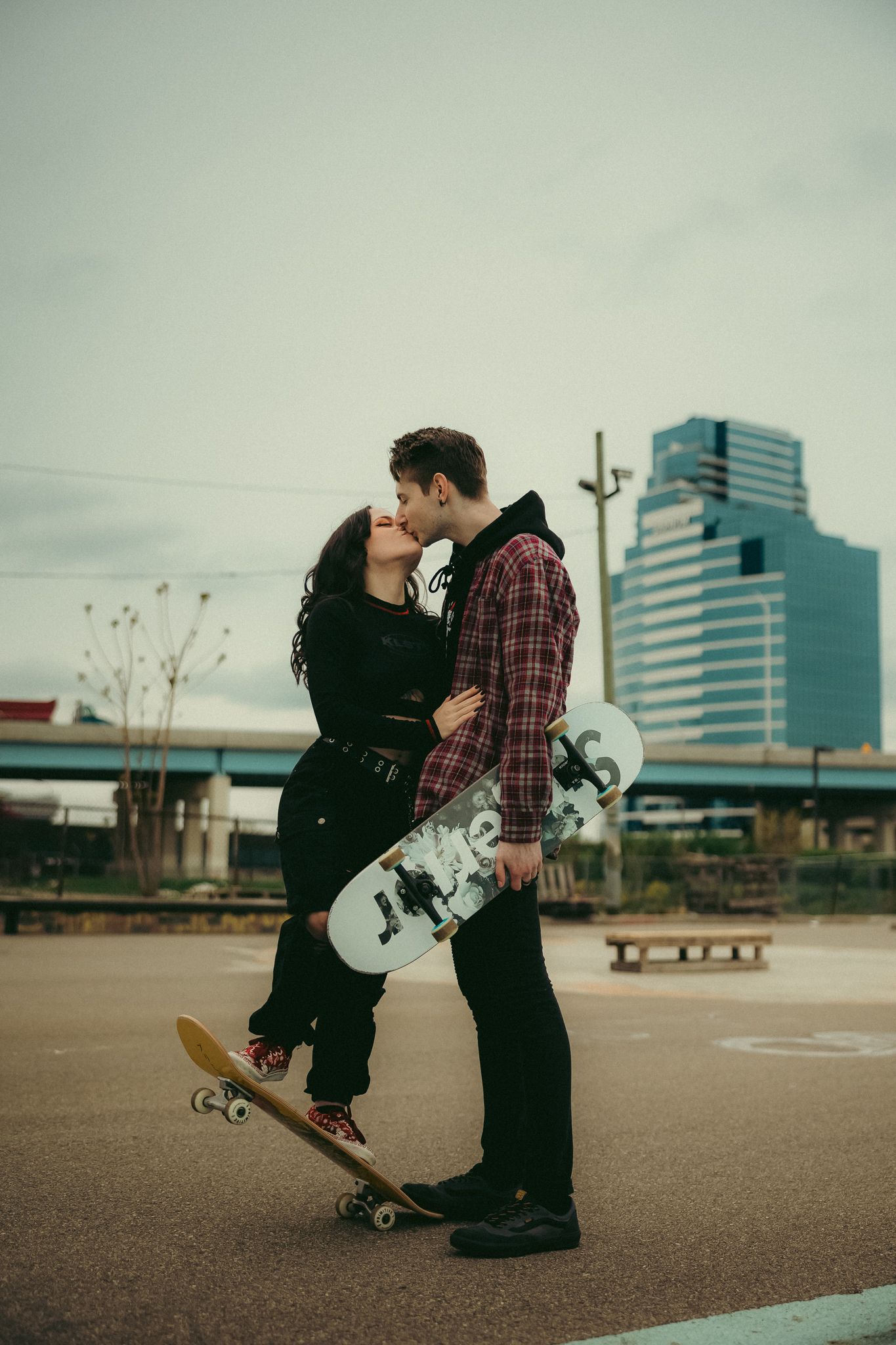 A skateboarding couple shares a romantic moment in an urban setting with office buildings in the background.