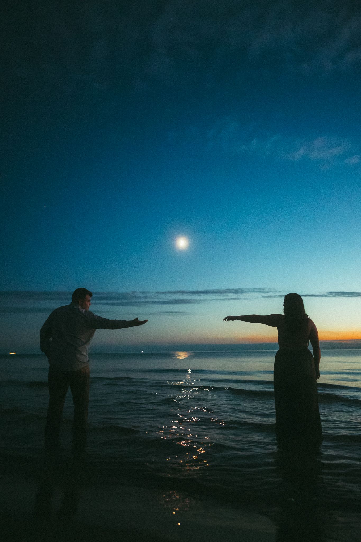 Silhouettes of people splashing water at sunset on a beach with a dramatic blue sky and moon in the background.